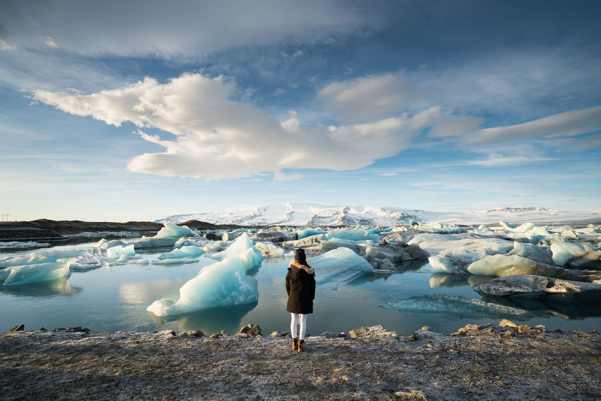 Person standing by a glacier lagoon with floating icebergs, under a cloudy sky.