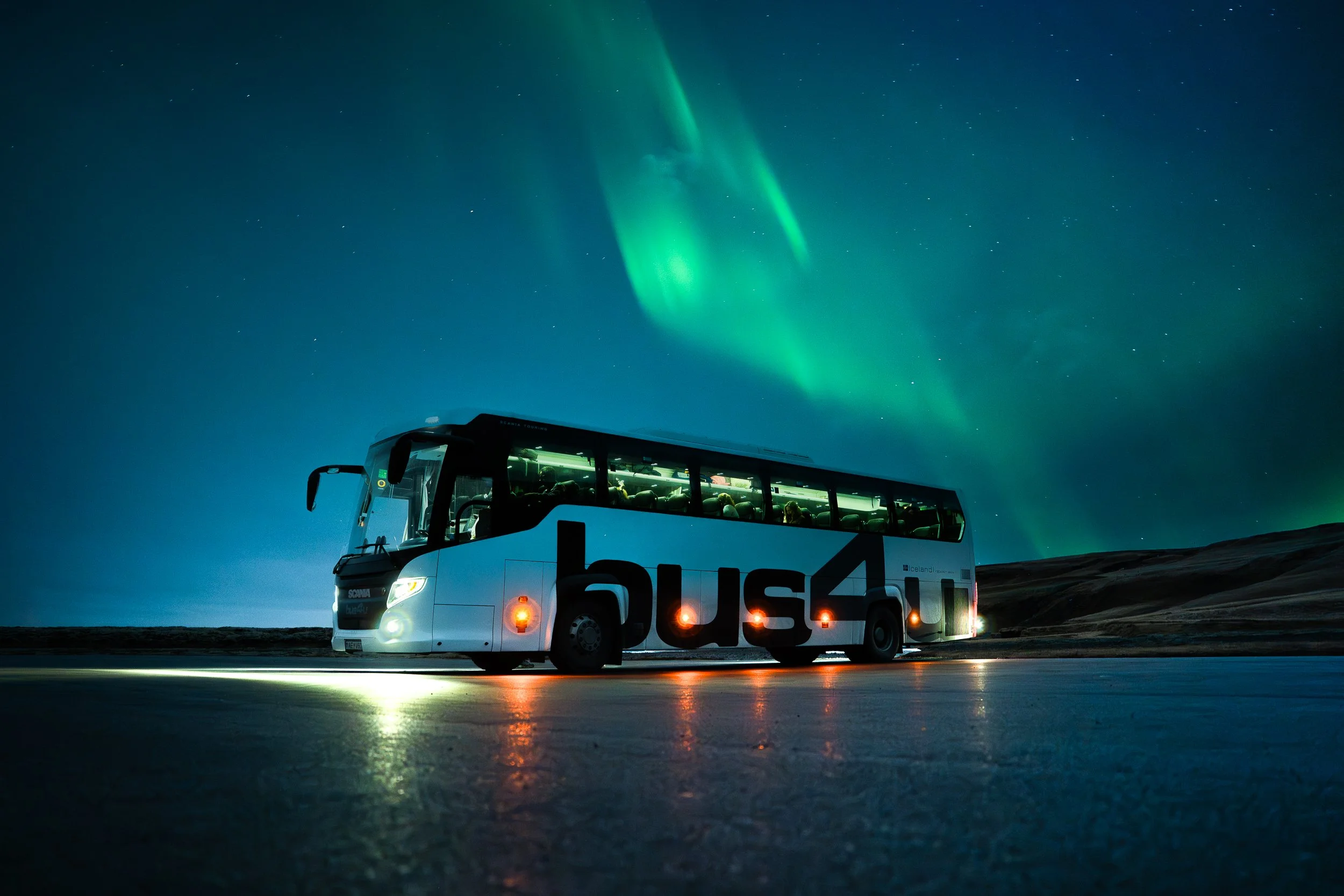 A bus with the logo 'bus4' on its side is traveling on an icy road at night under a sky illuminated by the Northern Lights.