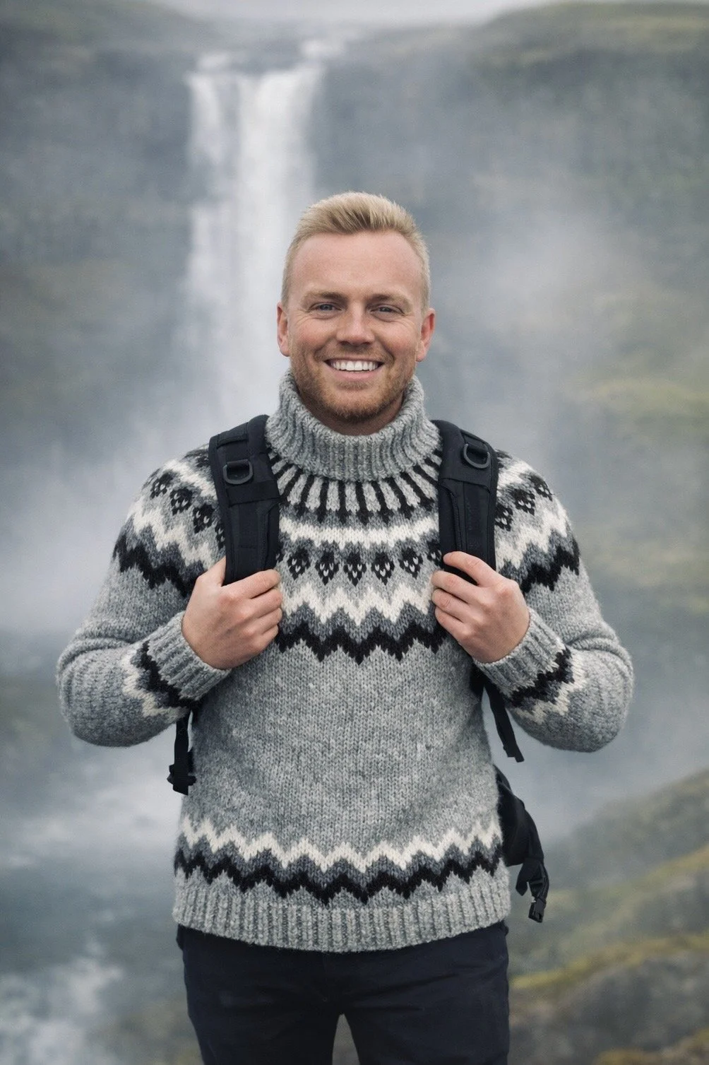 A man smiling outdoors in front of a waterfall, carrying a backpack, wearing a gray patterned sweater.