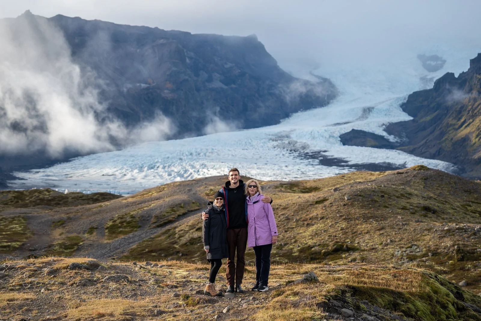 Three people standing together in front of a glacier and mountains, smiling, with one person's arm around another, outdoors.