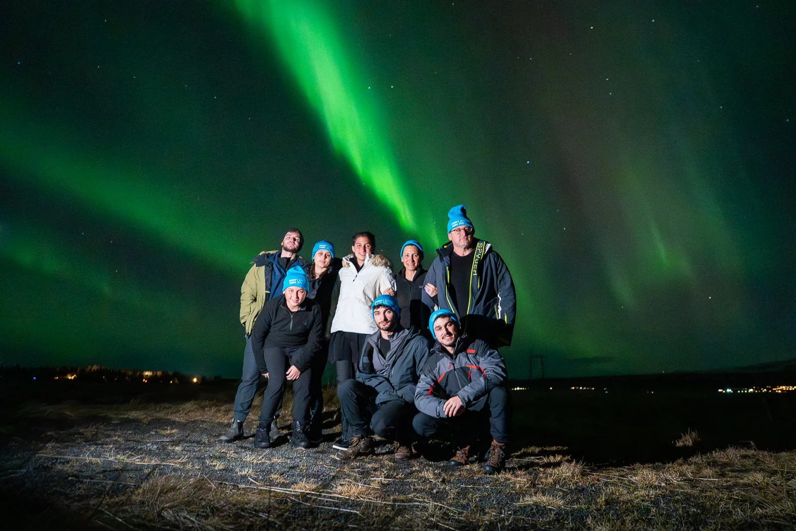 Group of seven people standing outdoors at night under the Northern Lights. They are dressed in winter clothing and wearing blue beanies.