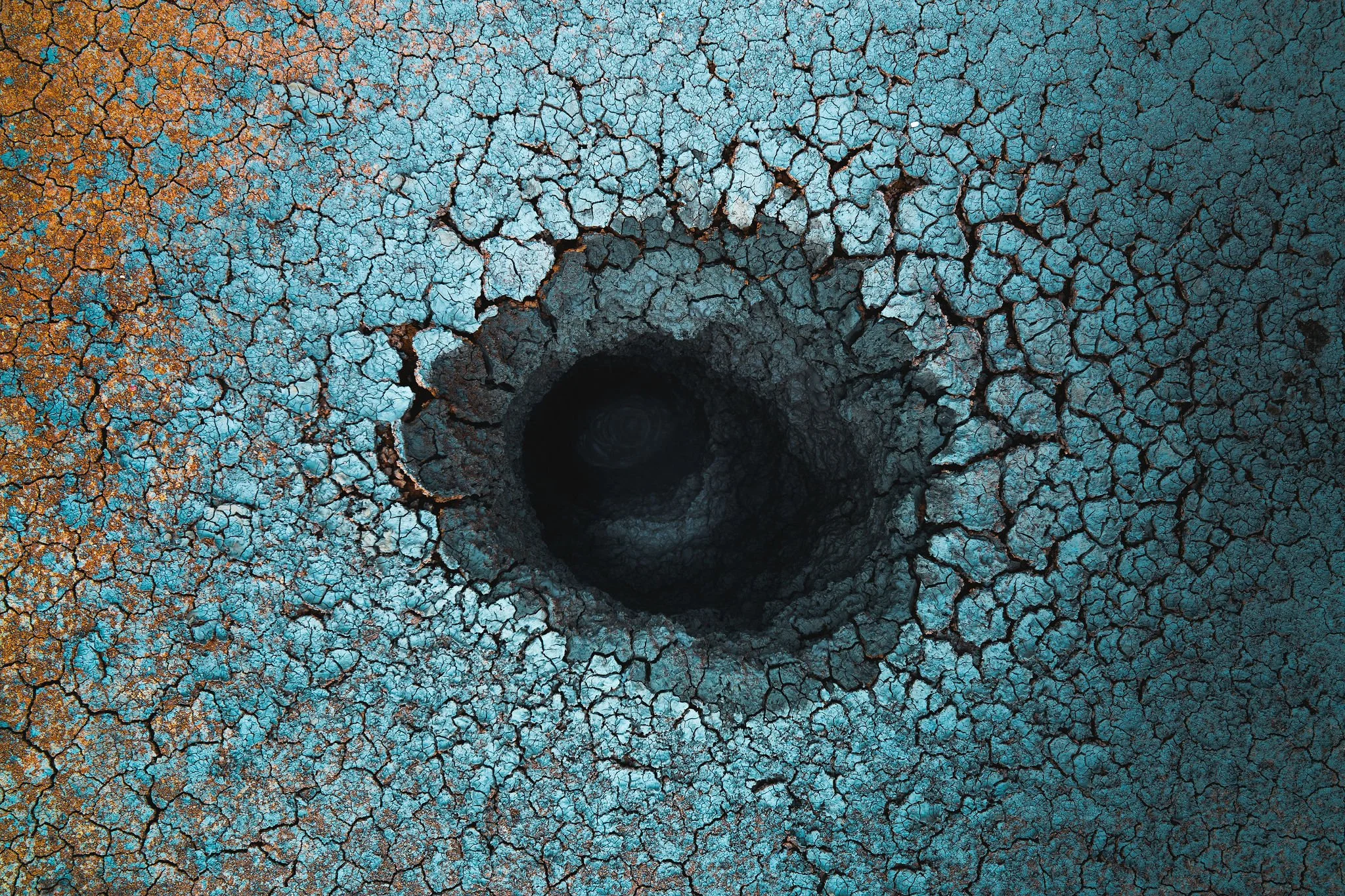 Aerial view of a dry, cracked earth surface with a deep dark hole in the center, showing contrasting blue and brown tones.