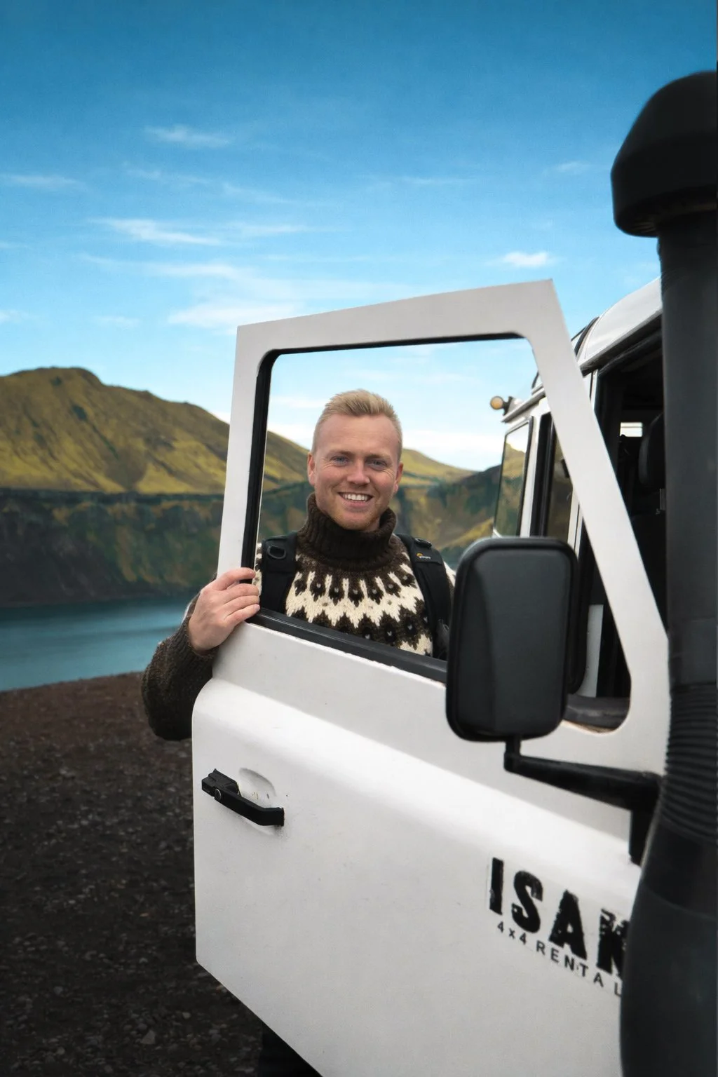 A smiling man standing in a white vehicle with an open door, with a scenic landscape of mountains and a lake in the background.