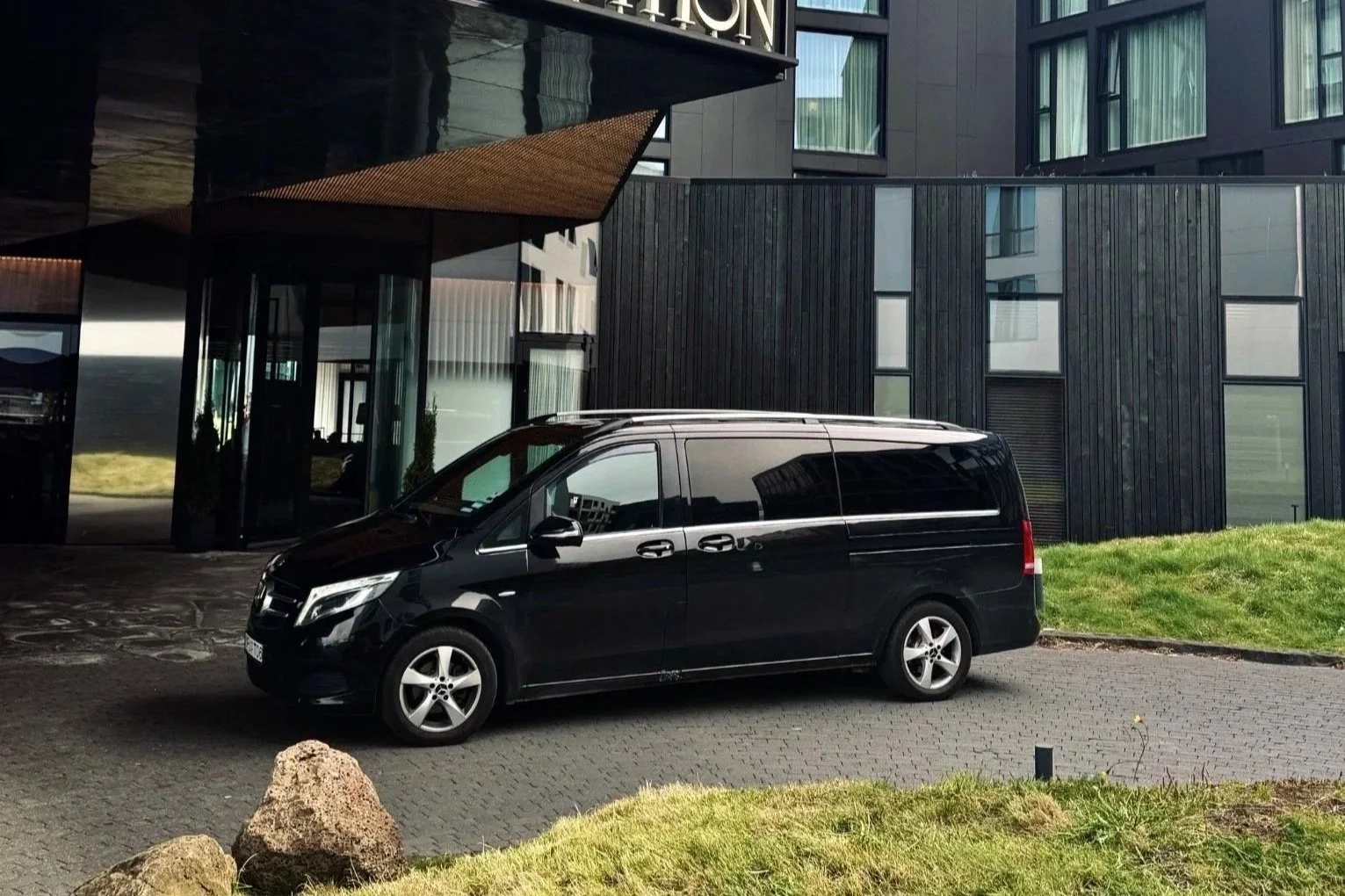 Black van parked outside modern building with glass windows and dark facade. Green grass and rocks in the foreground.