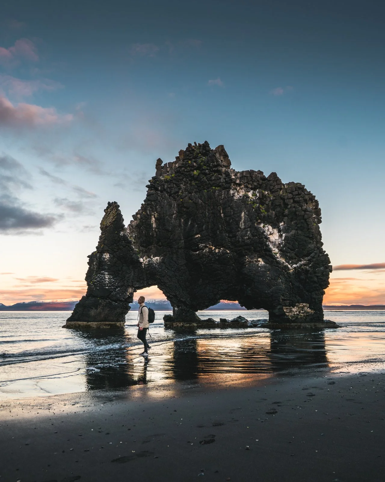 A person walking on a beach near a large rock formation that has natural arches, during sunset.