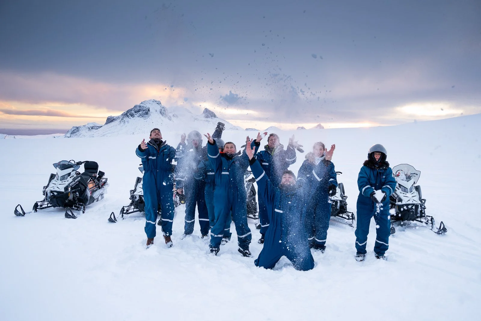 Group of people in blue winter suits playing with snow outdoors, with snowmobiles behind them and mountains in the distance, during sunset.
