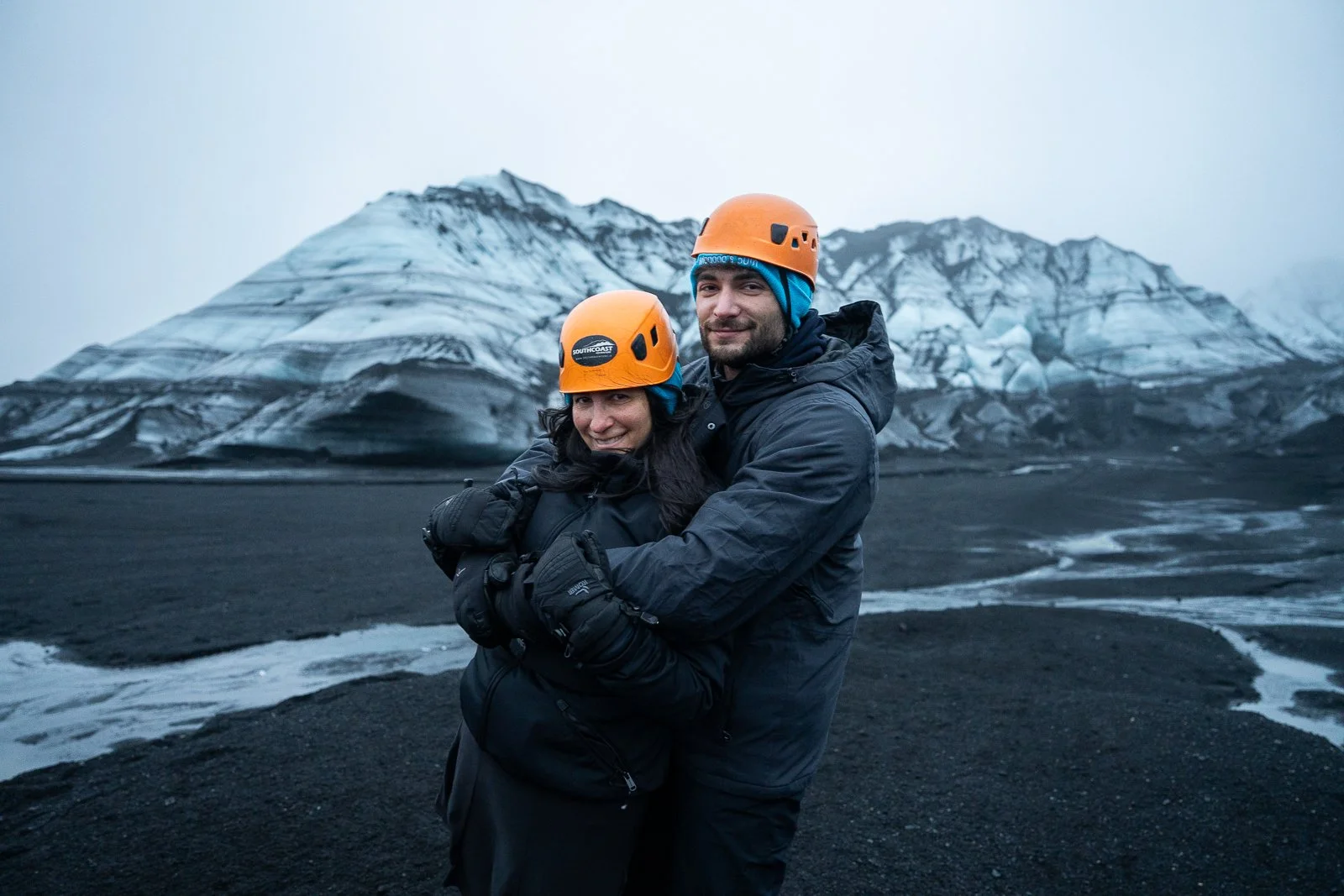 A couple wearing orange helmets and black outdoor gear hugging each other on a volcanic black sand landscape with snowy mountains in the background.