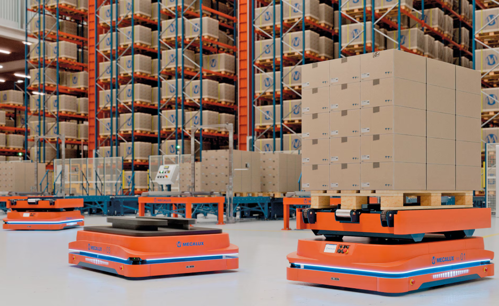 Autonomous orange warehouse robots moving pallets in a large storage facility with tall shelves filled with boxes.