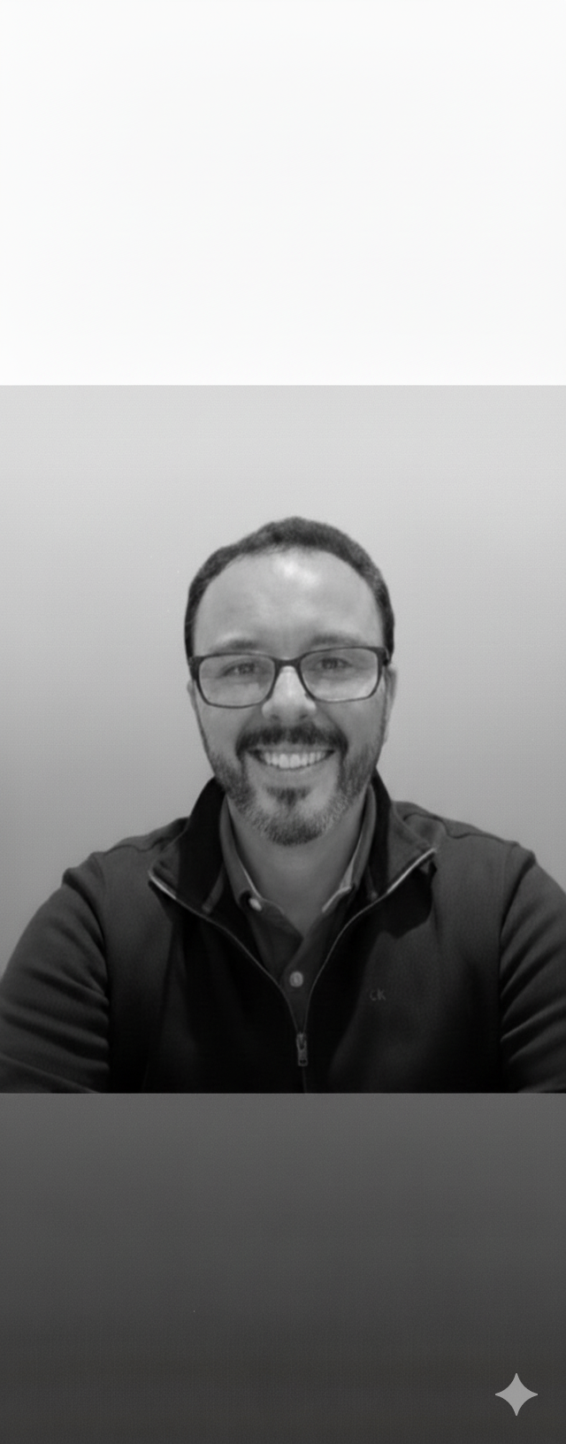 A man with glasses and a beard smiling for a selfie, wearing a dark jacket over a collared shirt, against a plain wall.