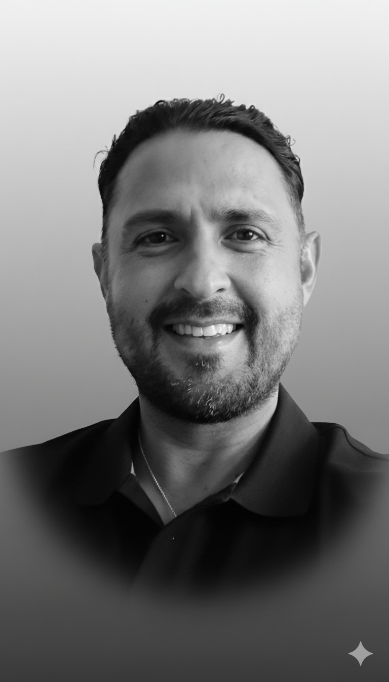 Black and white portrait of a smiling man with dark hair and a beard, wearing a dark collared shirt, against a plain light background.