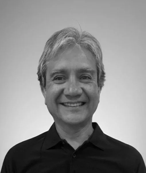 Black-and-white photo of a smiling man with gray hair, wearing a dark collared shirt, against a plain light background.