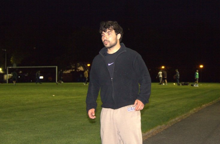 A man standing on a lit grass field at night, with people in the background, possibly a park or sports field.