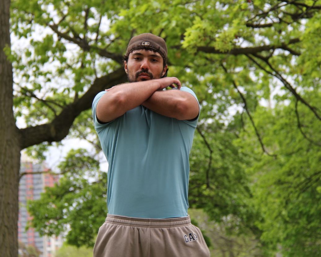 Young man standing outdoors in front of green trees, crossing his arms and looking at the camera.