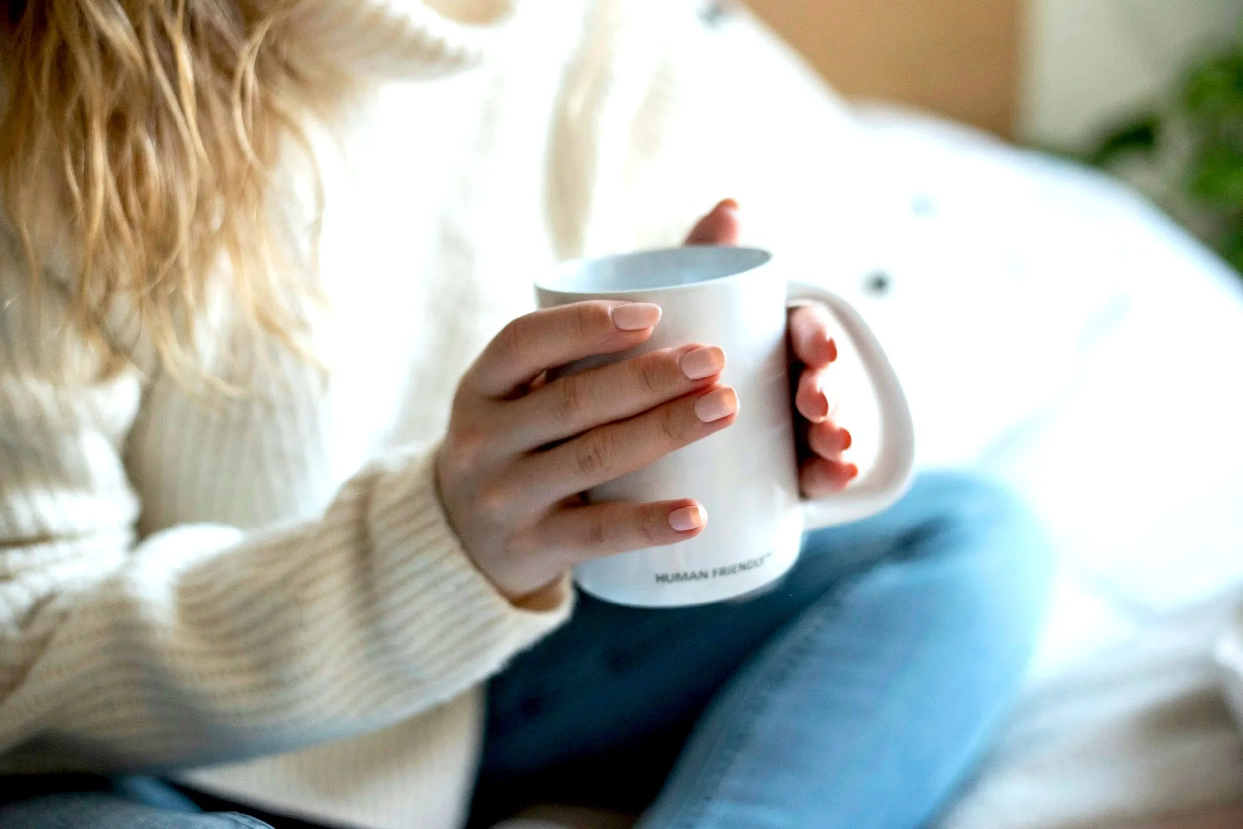 A woman with light skin and long blonde hair holding a white ceramic mug with text printed on it. She is wearing a cream-colored sweater and blue jeans, sitting on a blue surface. The background includes a green plant and light-colored furniture.