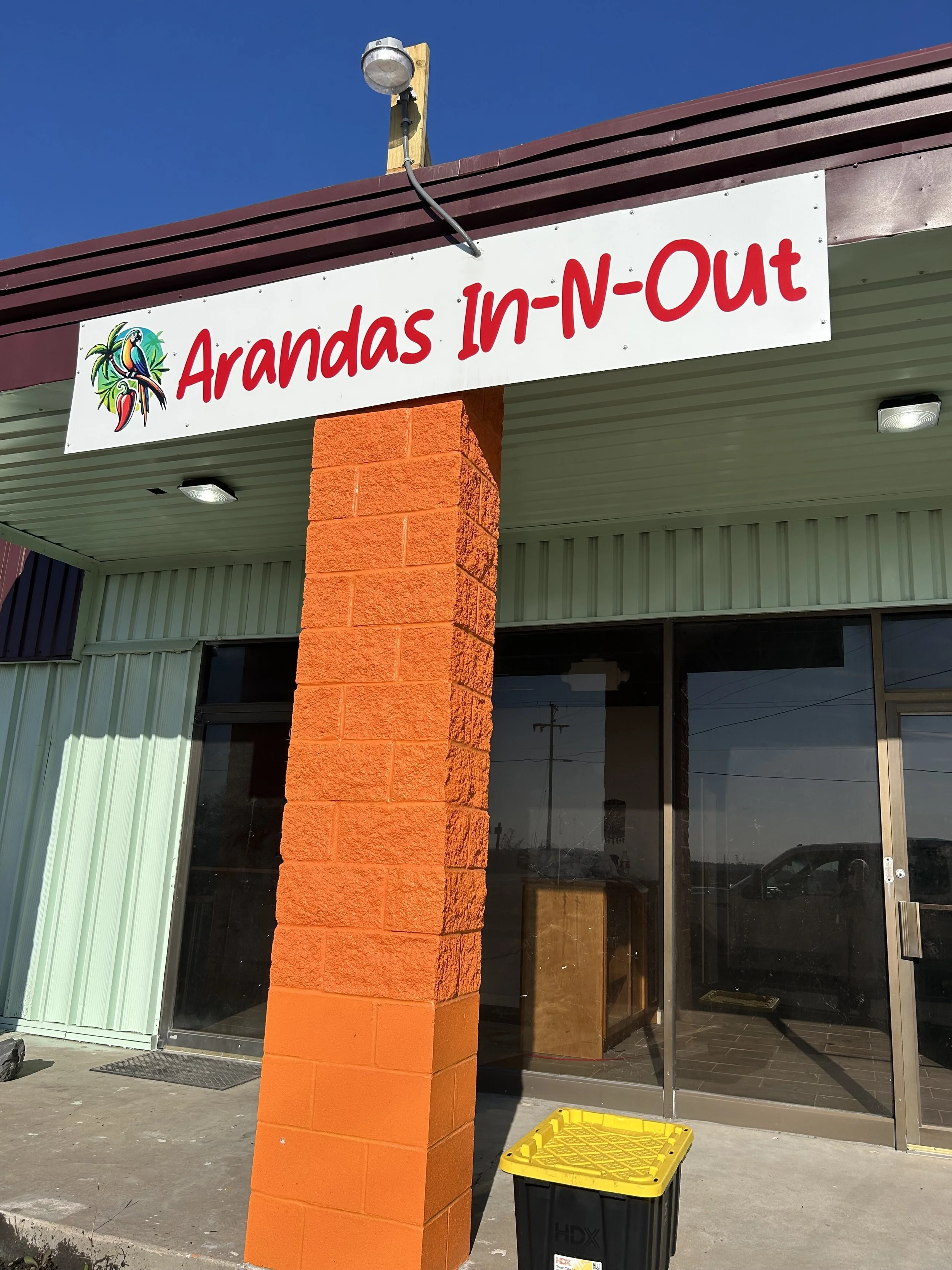 Storefront with a sign that reads 'Arandas In-N-Out', featuring a colorful parrot logo, an orange brick column, glass doors, and a yellow and black trash bin outside.