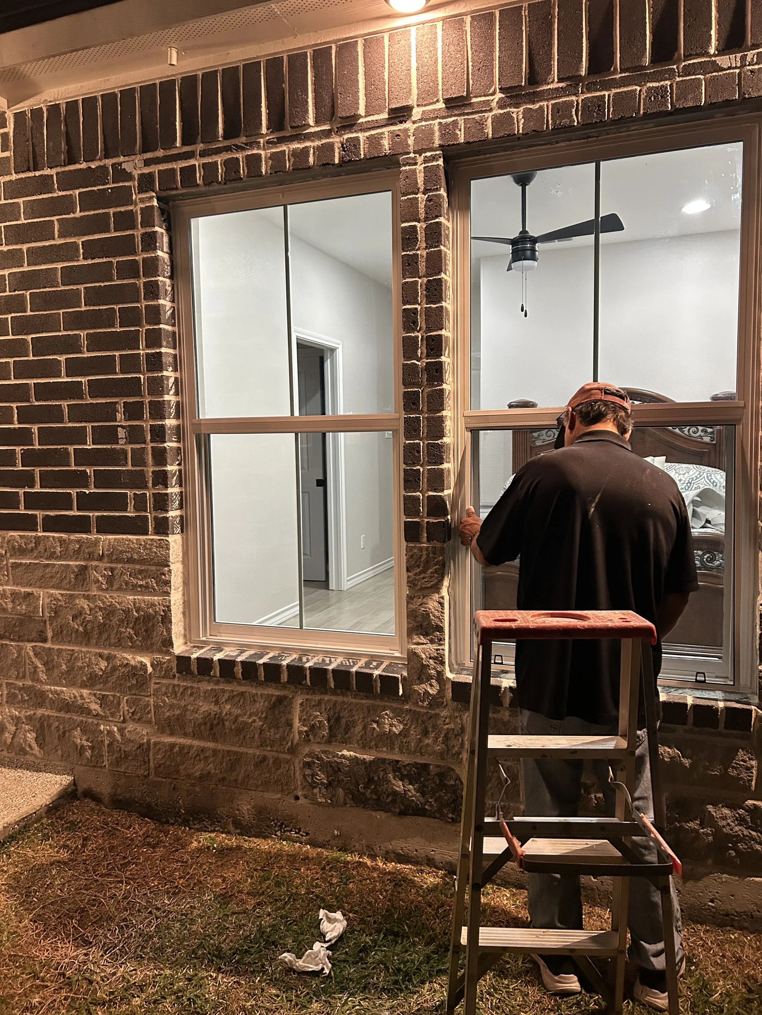 Man installing or repairing window on brick house exterior at night, using a ladder