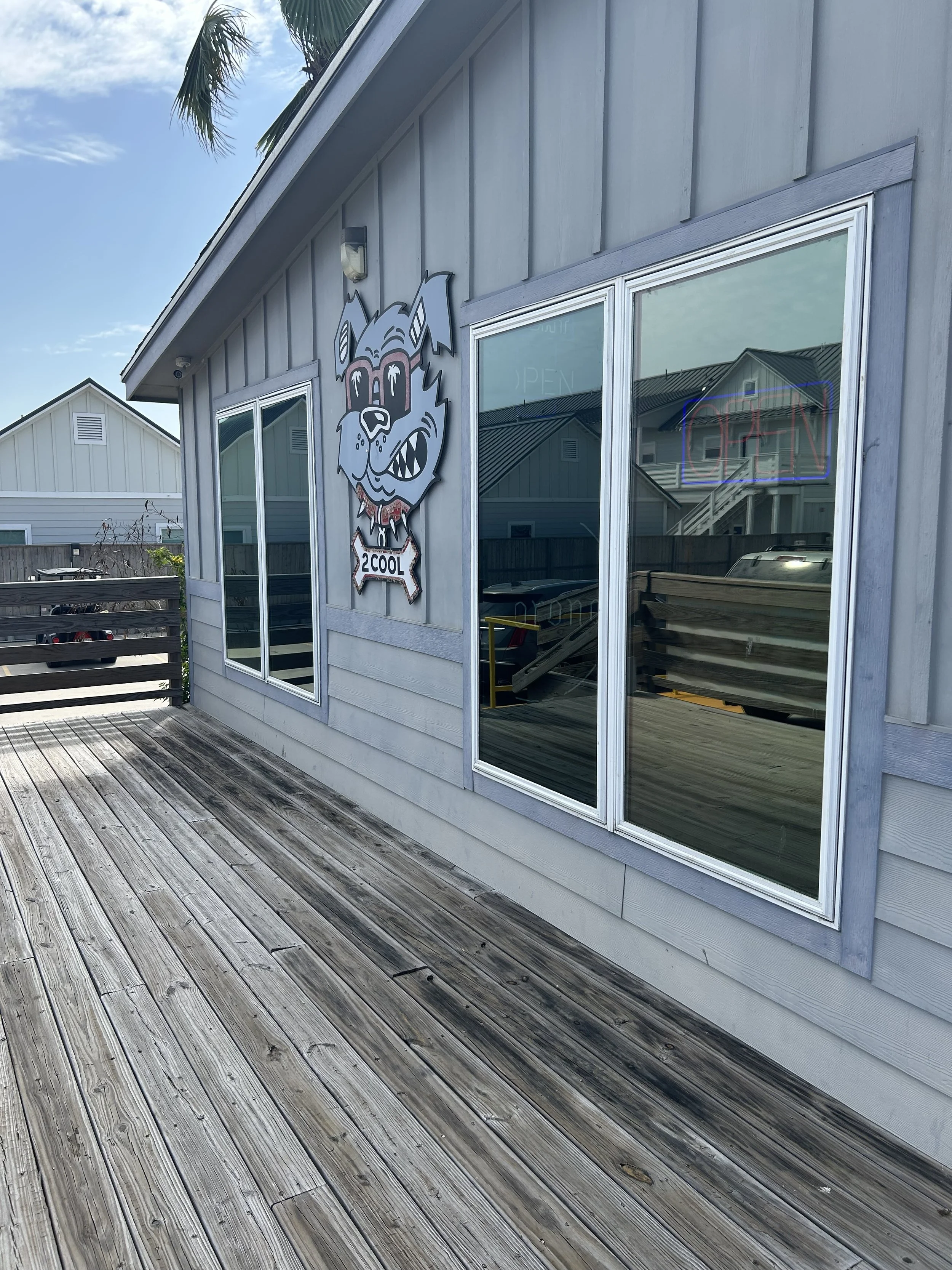 The exterior of a small building with large windows and a wooden deck. There is a cartoonish dog sign with a bowtie and the words '2COOL' hanging on the wall, and a neon 'OPEN' sign in one of the windows.