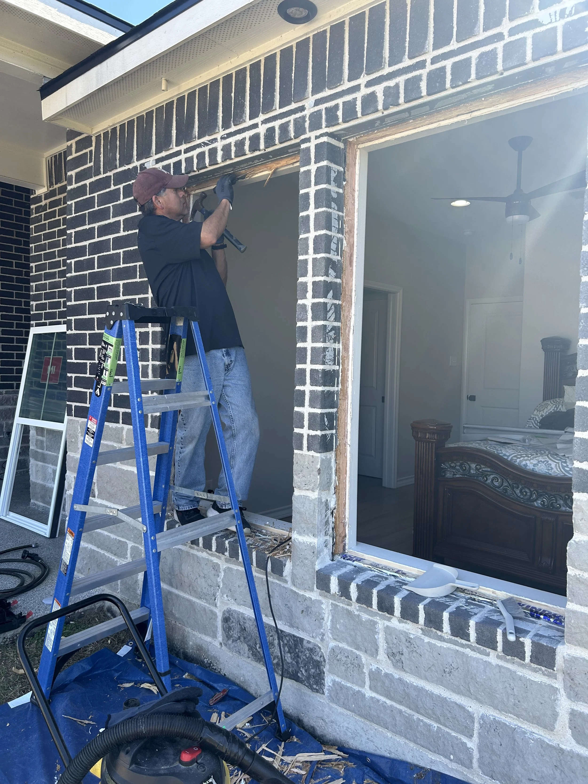 A man standing on a blue ladder working on the exterior brick wall of a house, using a tool to remove or install the framing around a large window opening, with construction tools and materials nearby.