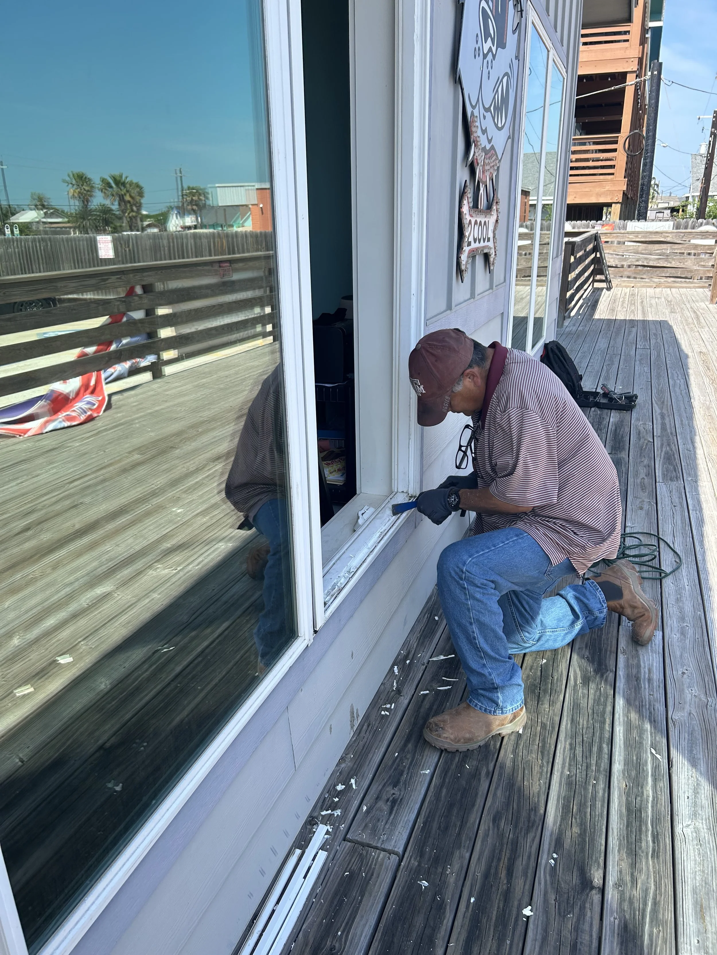 A man kneeling on a wooden deck, working on the bottom window frame of a building, wearing a maroon cap, striped shirt, jeans, and brown boots.