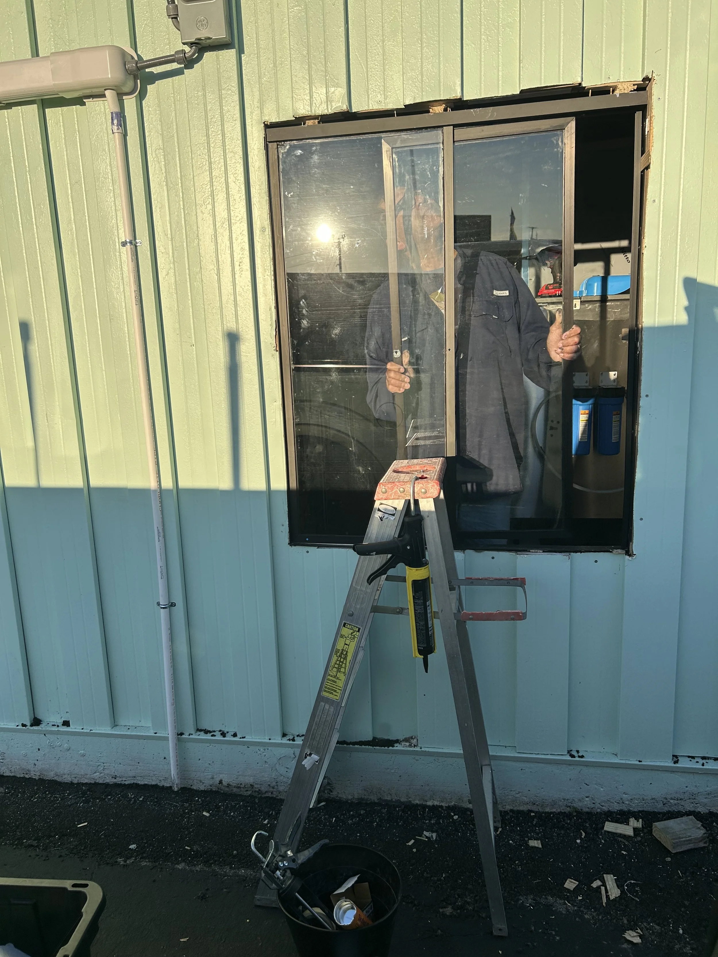 Person installing a window in a green metal building, with a ladder, tools, and debris on the ground.