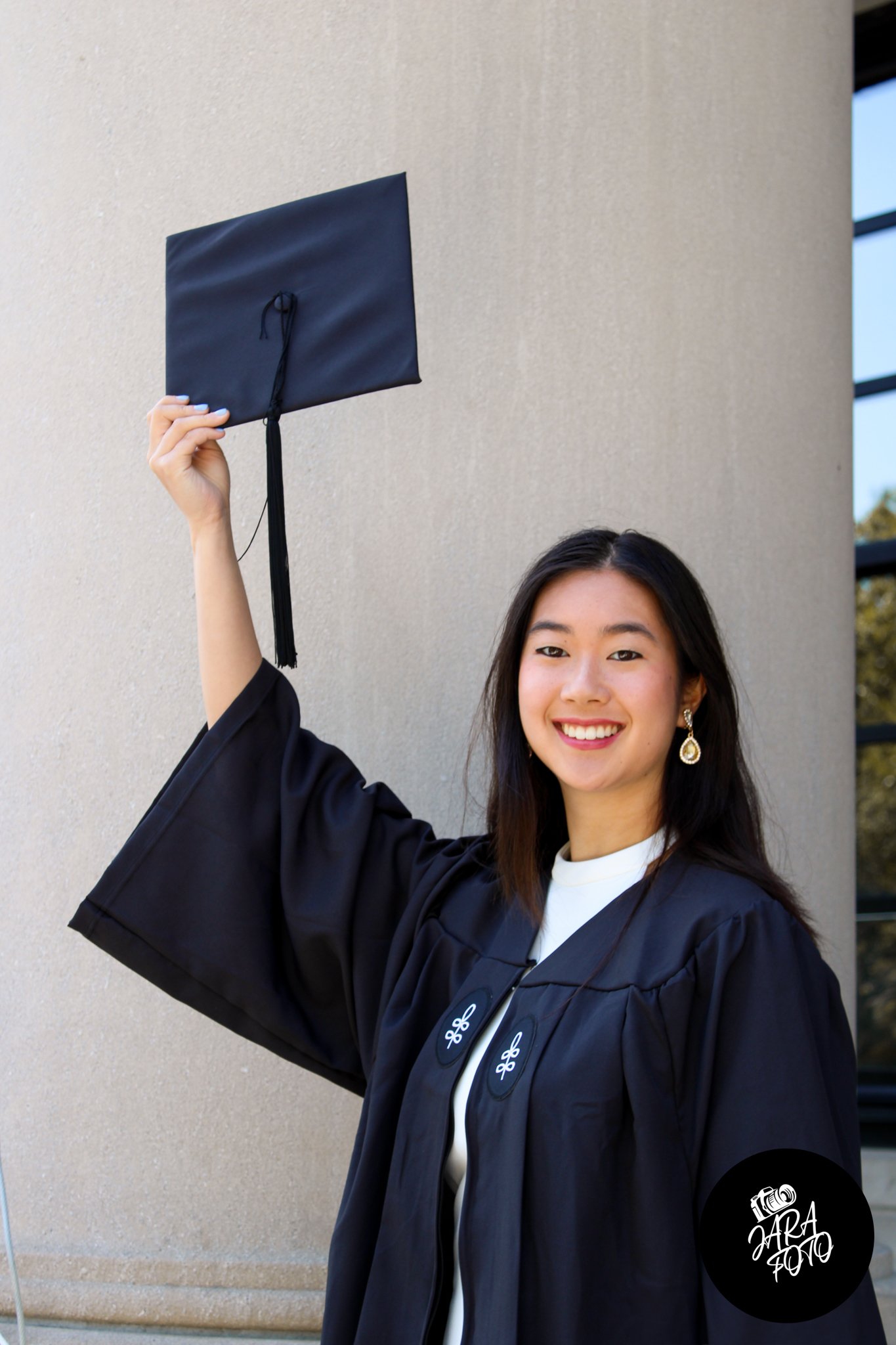 A young woman wearing a graduation gown holds up a mortarboard cap while smiling, standing in front of a concrete column.