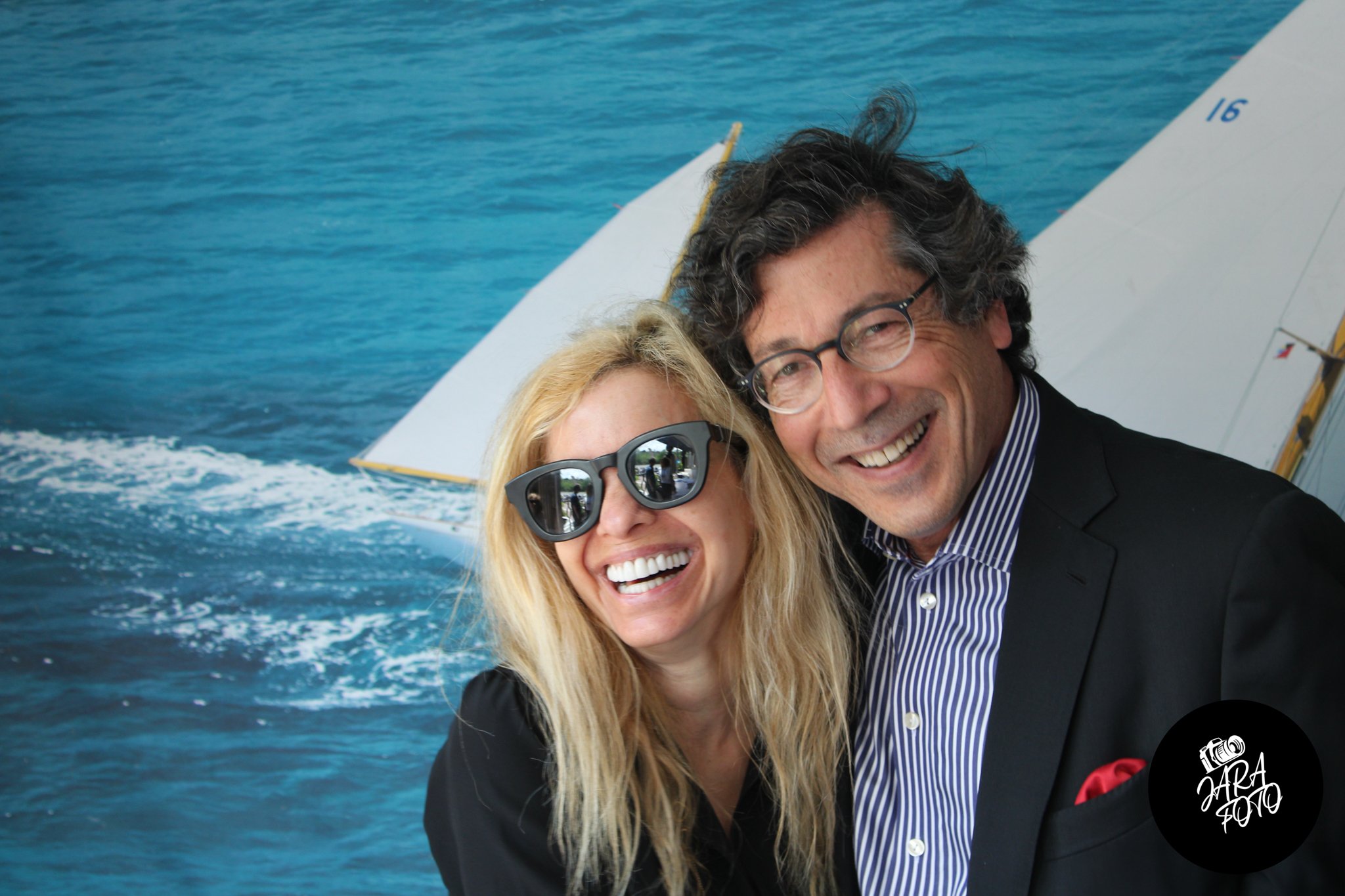 A smiling woman with blonde hair and sunglasses stands next to a man with glasses, in front of a backdrop featuring a sailboat on the ocean.