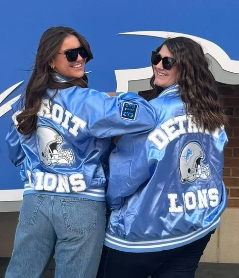 Two women wearing Detroit Lions satin jackets and sunglasses, smiling and posing back-to-back in front of a blue wall with a lion graphic.