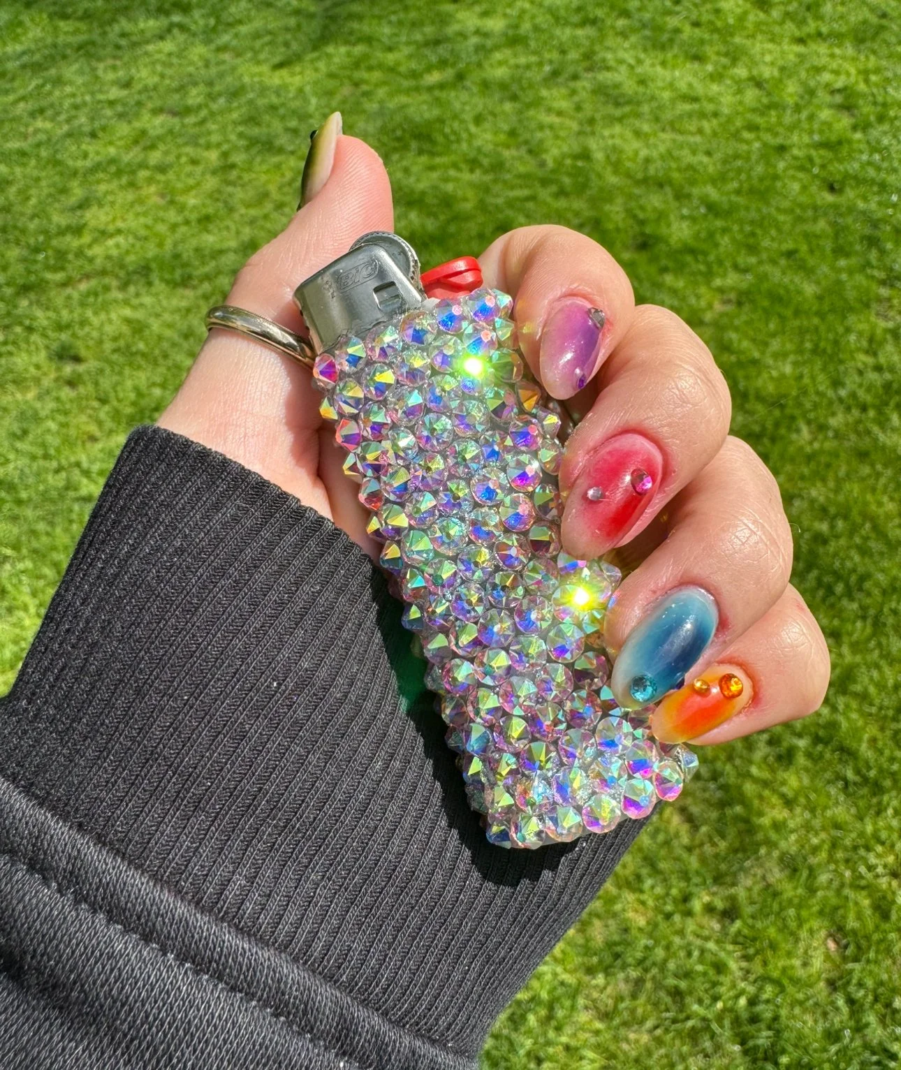 Hand with colorful pastel and jewel-toned painted nails holding a sparkly rhinestone-covered lighter outdoors on green grass.