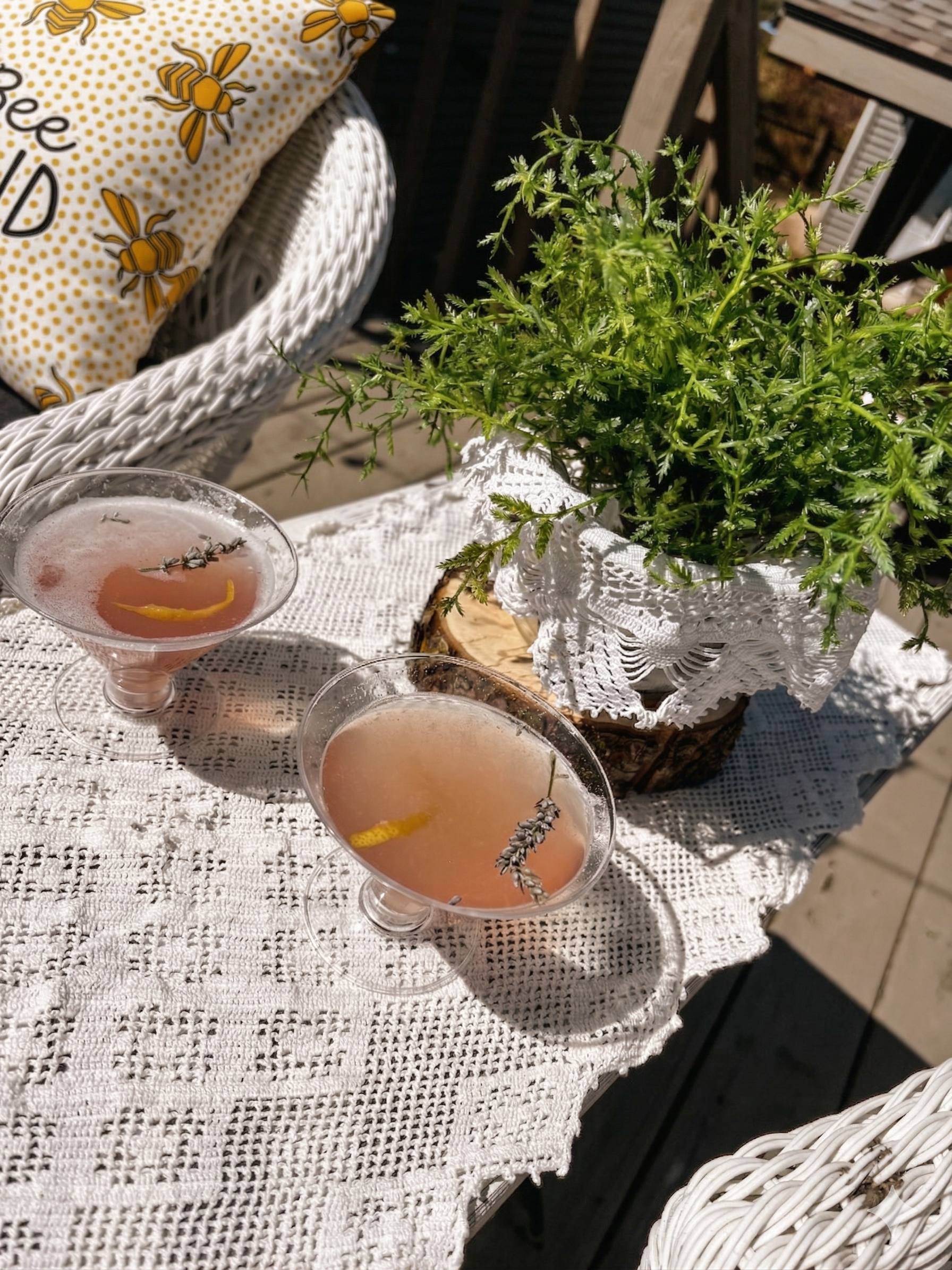 Two pale pink mocktails in clear stemmed coupe glasses with a sugared rim, a lemon twist, and a lavender sprig sitting on a white lace tablecloth with a potted green plant to the right of a white wicker chair.