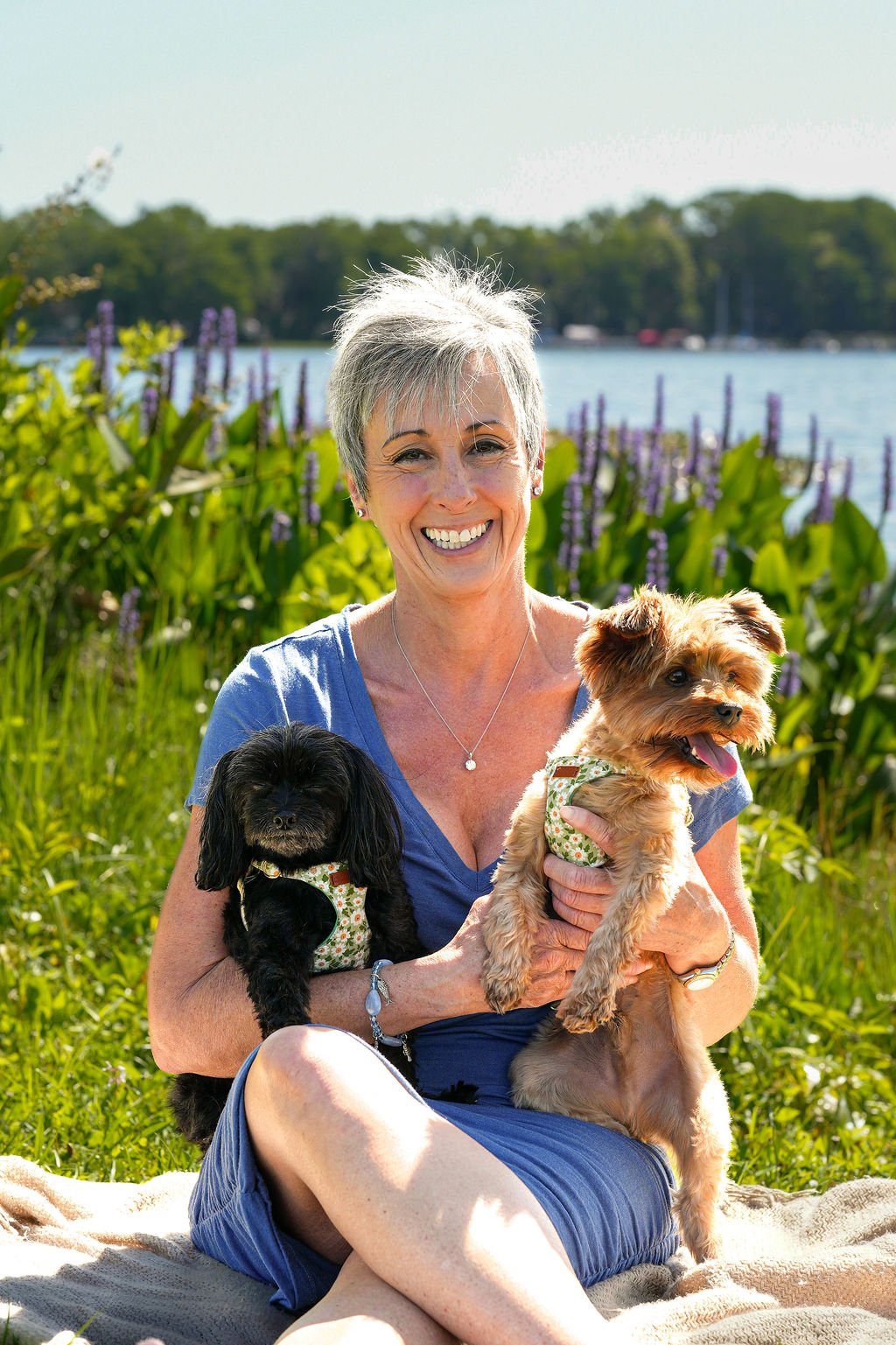 A woman with short gray hair smiling while holding two small dogs, one black and one tan, outdoors near a lake with purple and green plants in the background on a sunny day.