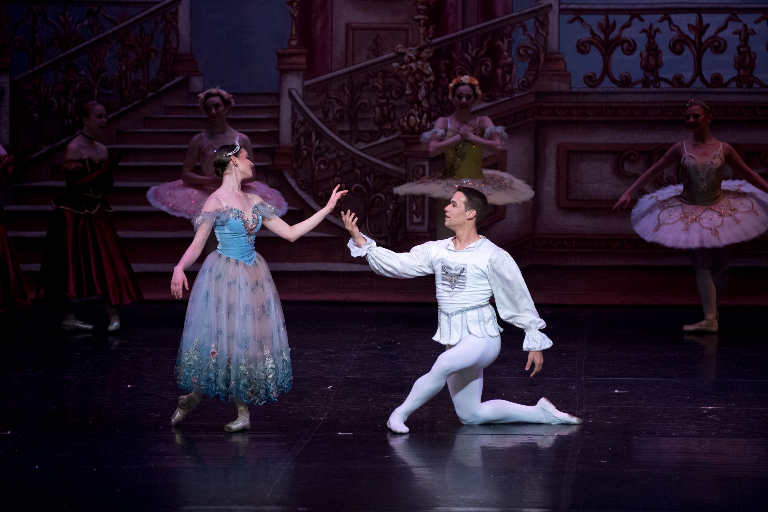 Ballet dancers performing on stage, with a male dancer on one knee reaching out to a female dancer in a blue and teal tutu, in front of a grand staircase backdrop with ornate railings.