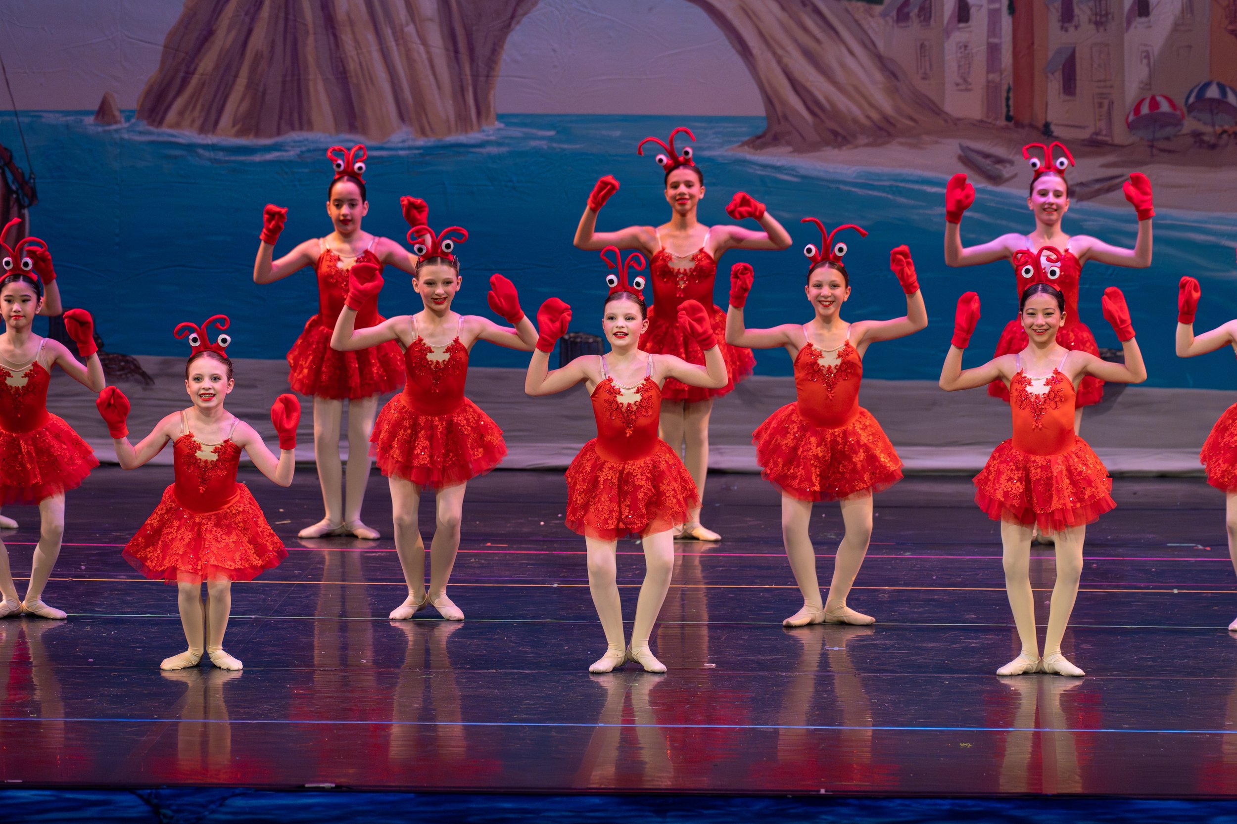 Children dressed in red costumes performing a dance on stage with a painted ocean and island backdrop.