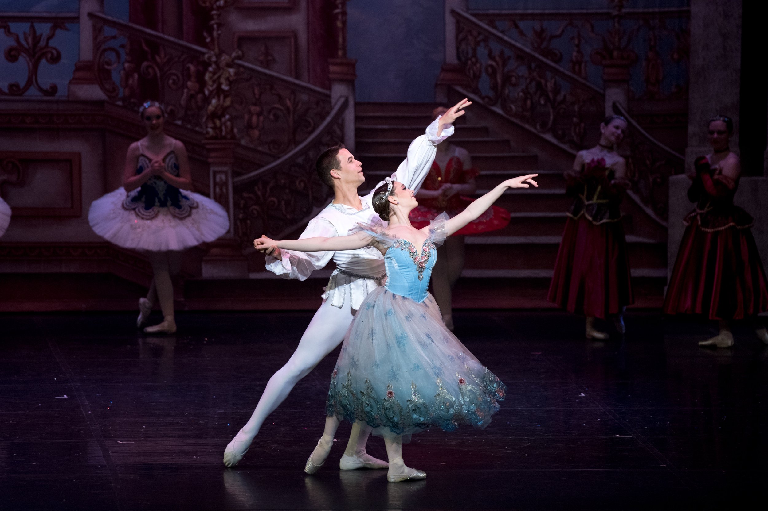 Two ballet dancers performing on stage with a backdrop of ornate stairs and other dancers in elegant costumes in the background.