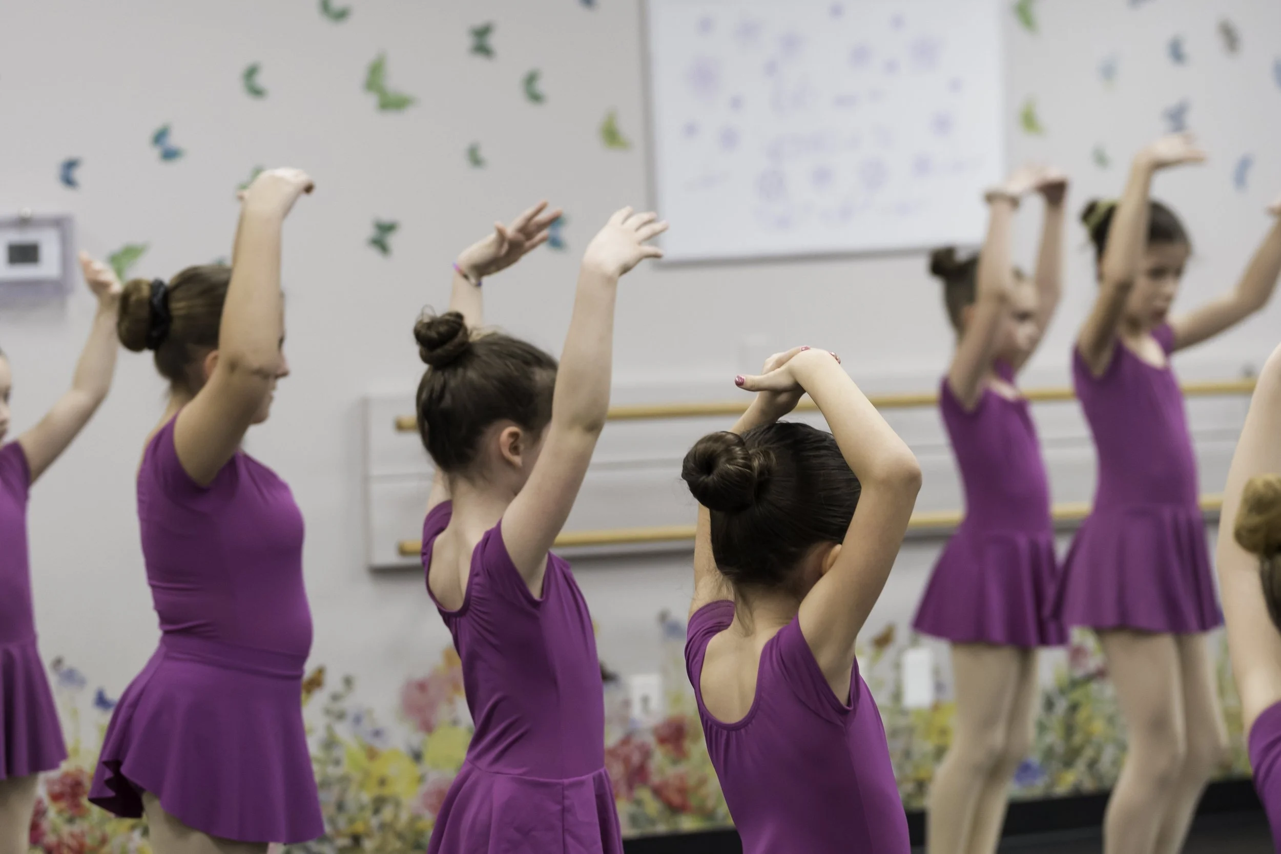 Group of young girls in purple ballet dresses practicing dance moves in a dance studio.