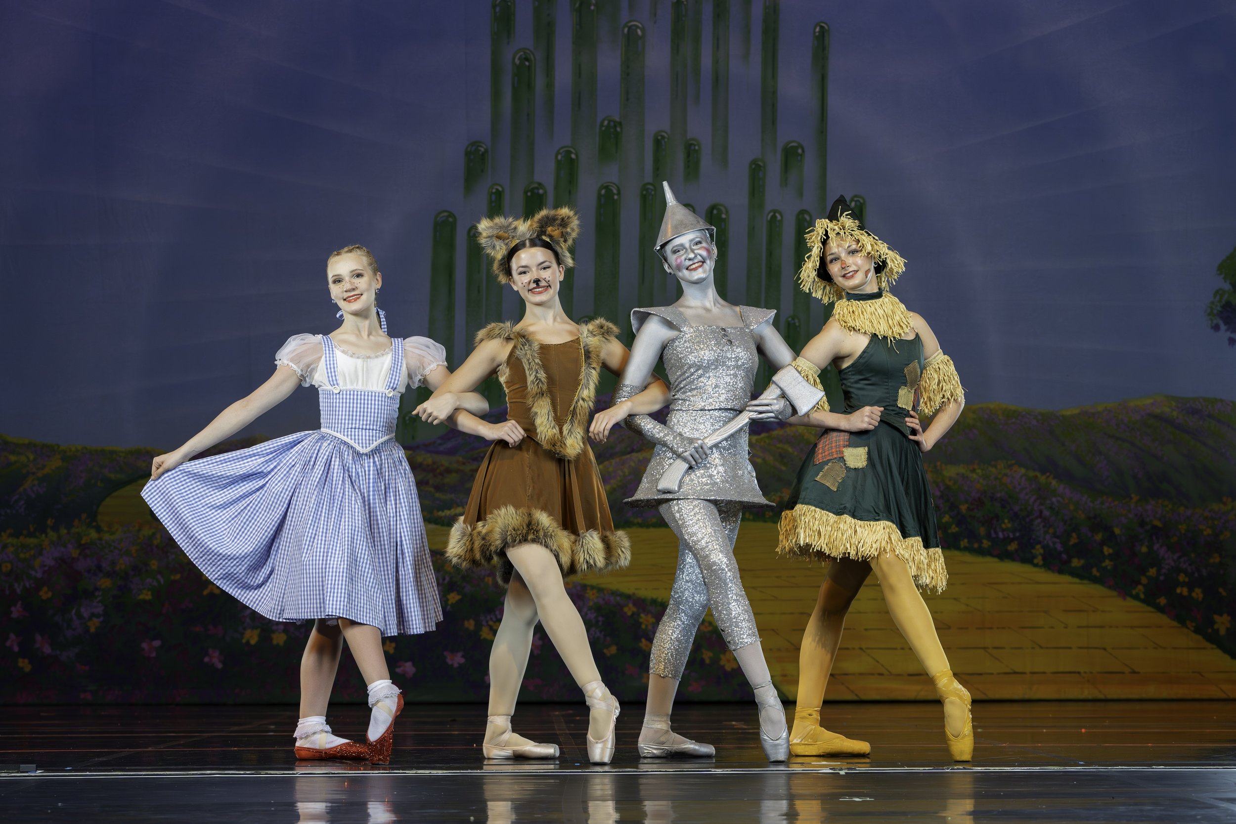 Four women dressed as characters from The Wizard of Oz, standing on stage with a colorful landscape backdrop, including a yellow brick road.