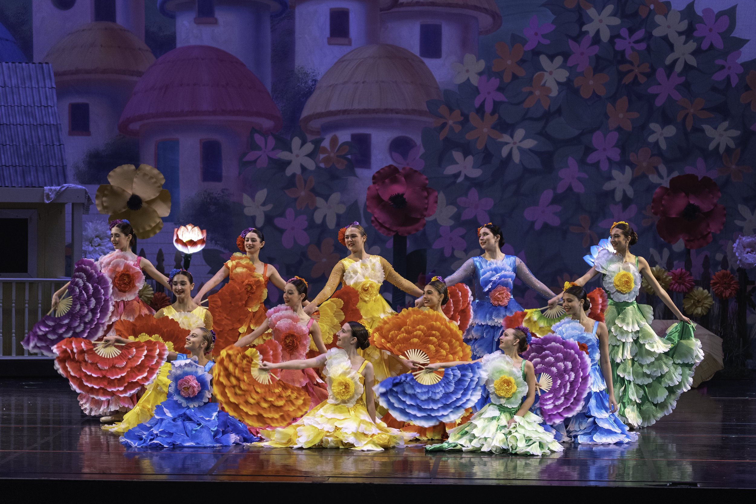 A group of dancers in colorful floral costumes performing on stage with a bright, whimsical background of flowers and fairy-tale buildings.