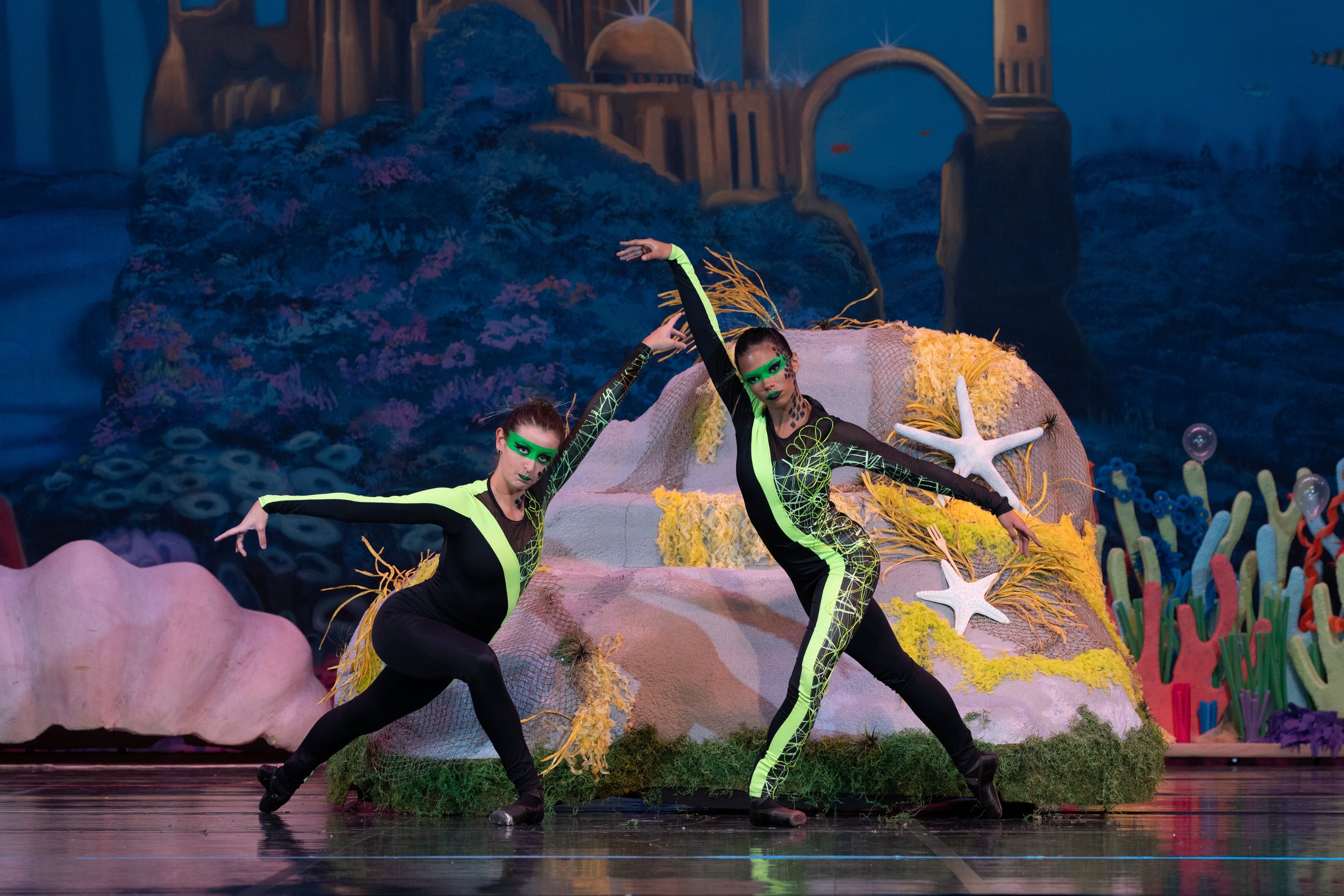 Two performers in black and neon green costumes pose on stage with a colorful ocean-themed backdrop and decorative elements resembling coral, starfish, and seaweed.
