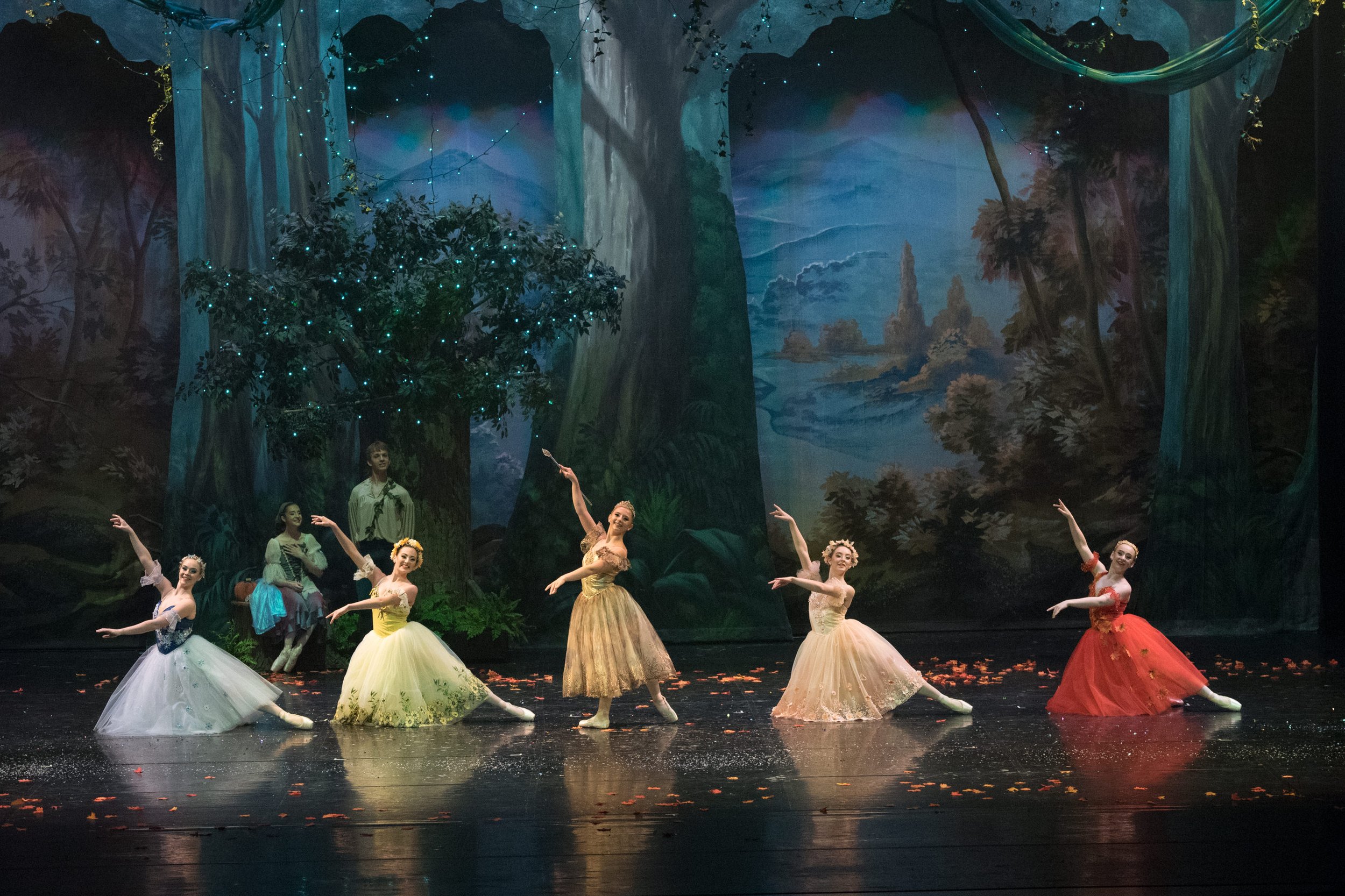 Ballet dancers performing on stage in a forest scene with trees and a river backdrop, decorated with lights and falling leaves.