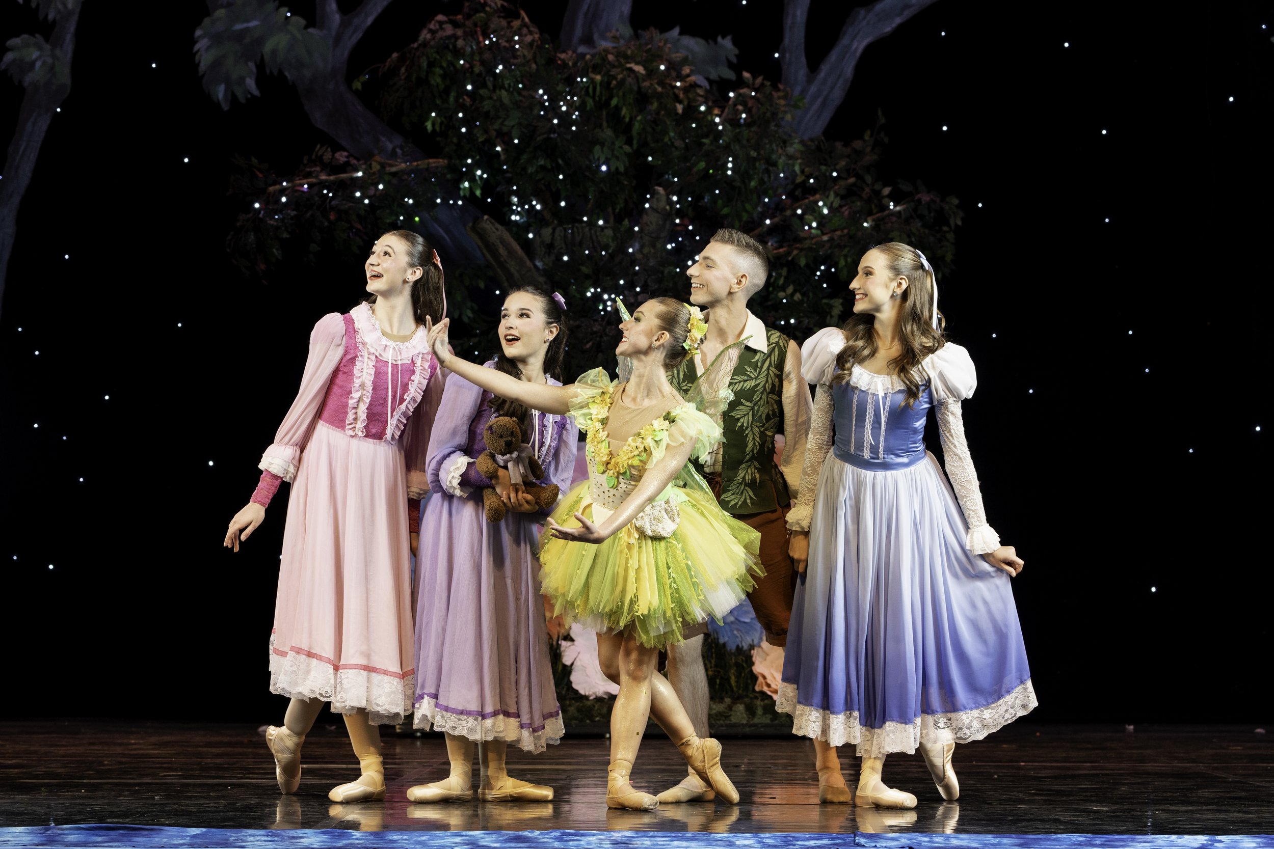 Ballet dancers performing on stage with a dark background and a large decorative tree with lights.
