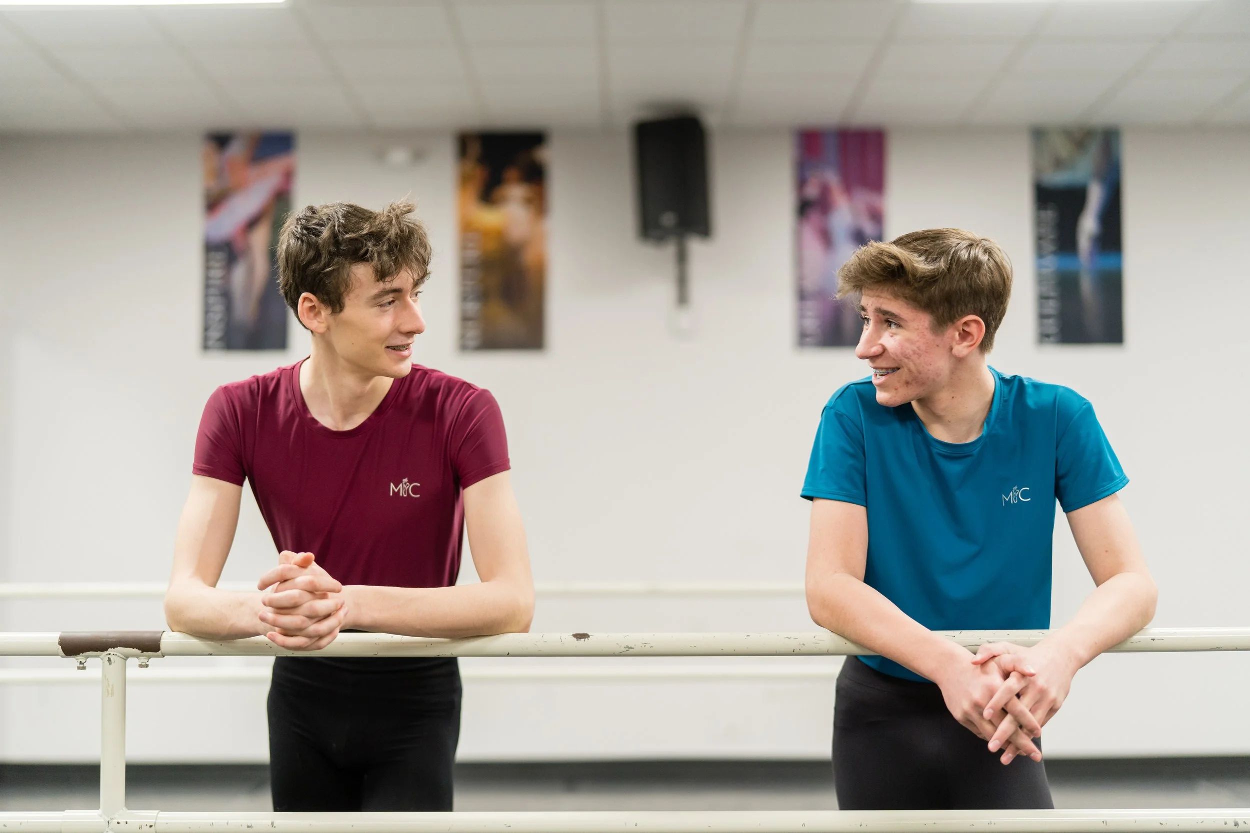Two young male figure skaters talking and smiling at each other, wearing athletic shirts, standing by a ballet bar in a training room.