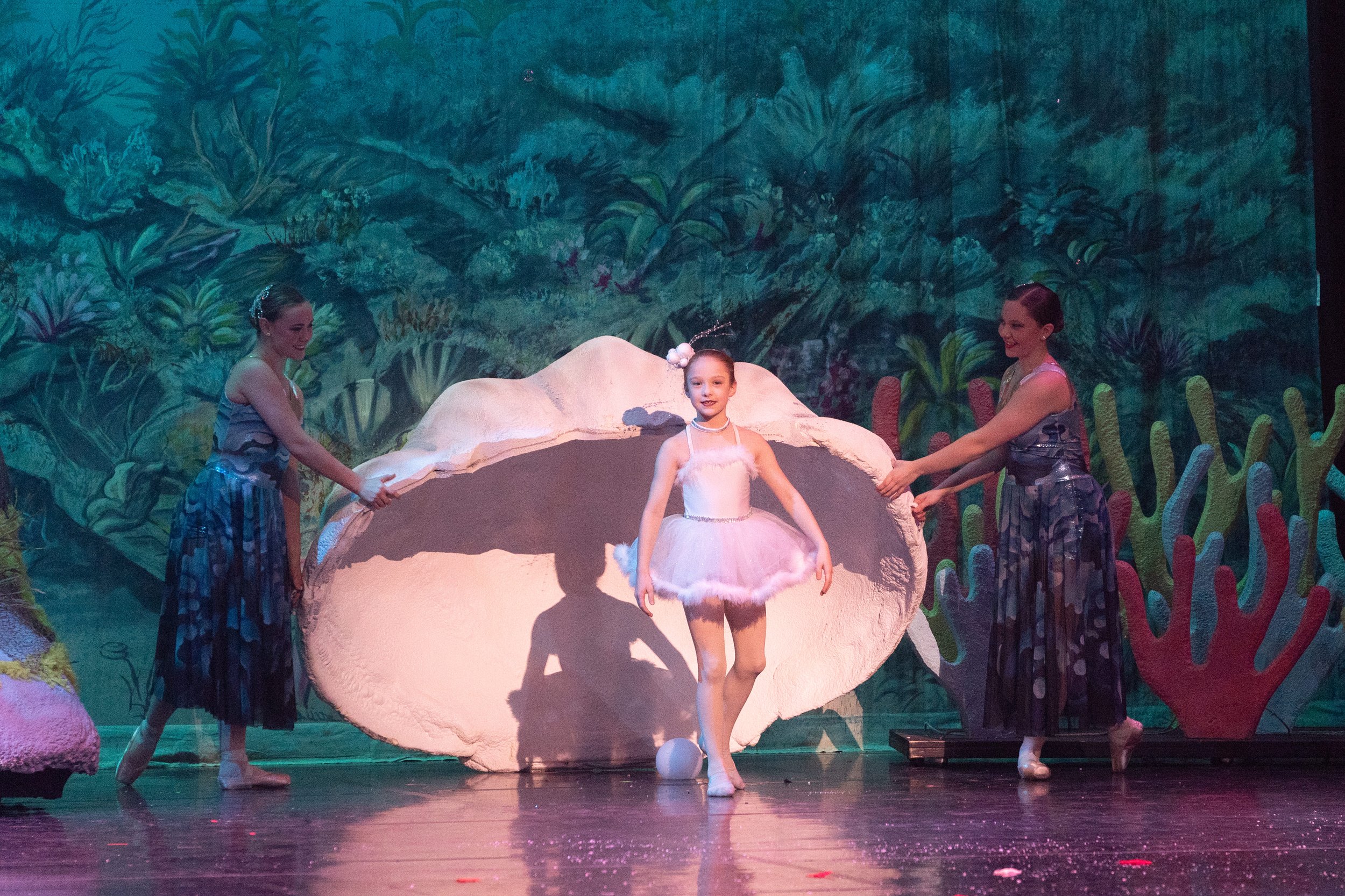 Two ballerinas hold open a giant clam shell while a young girl in a pink tutu dances in front of a colorful jungle backdrop.