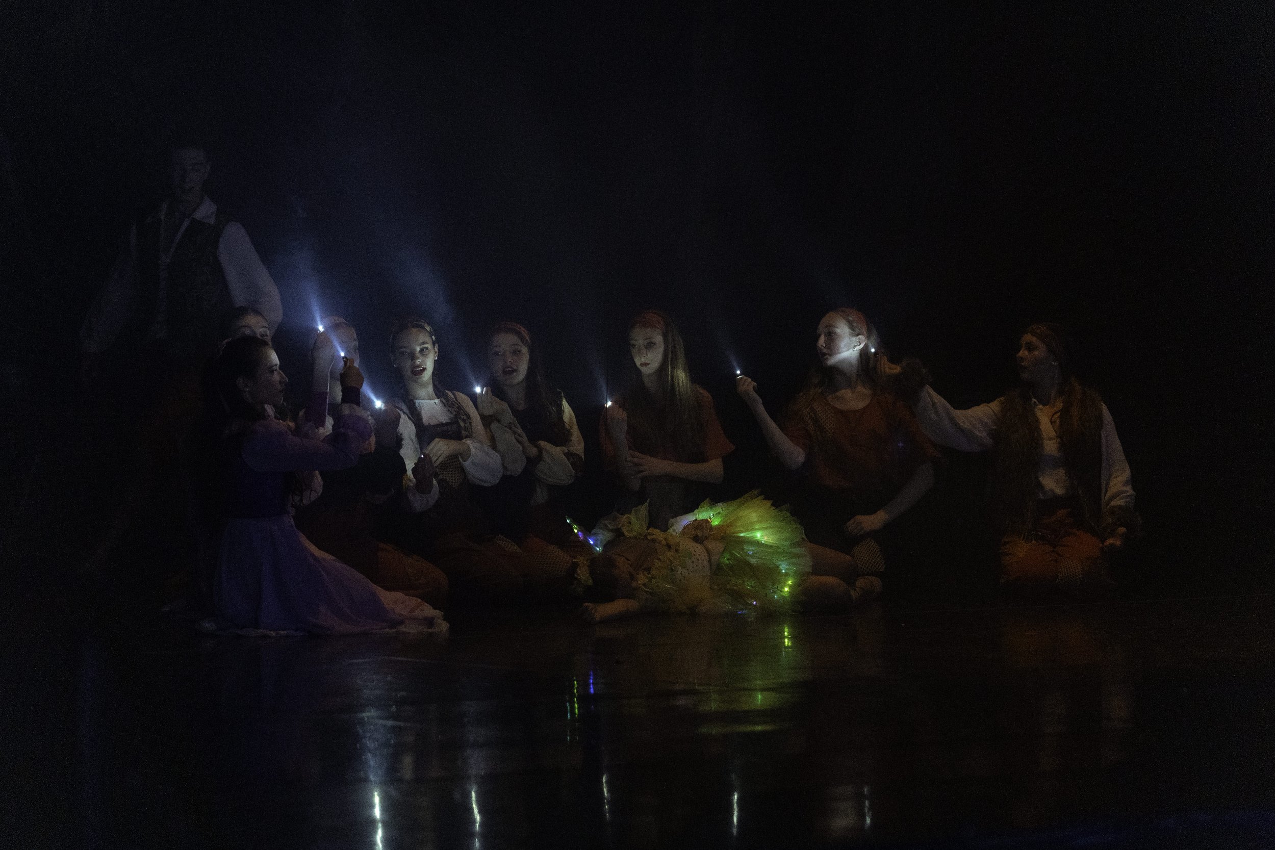 A group of girls dressed in costumes sitting in a dark room, holding sparklers, with some colorful glowing props on the floor.