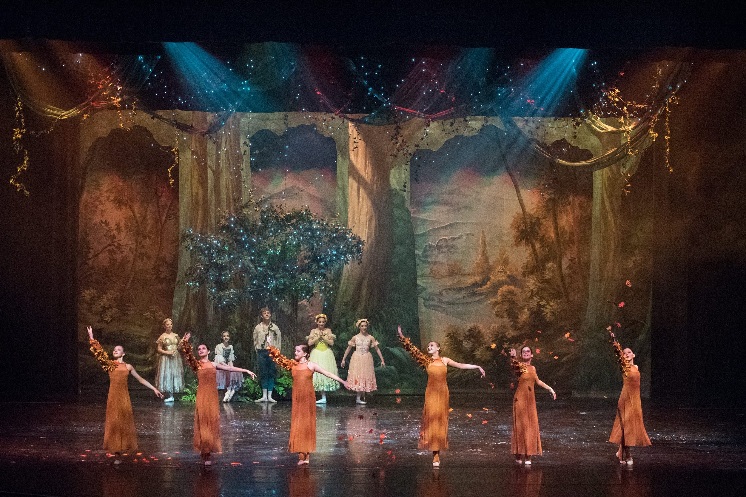 Ballet dancers performing on stage with a forest-themed backdrop, lit with blue and warm lighting, and dancers holding floral props.