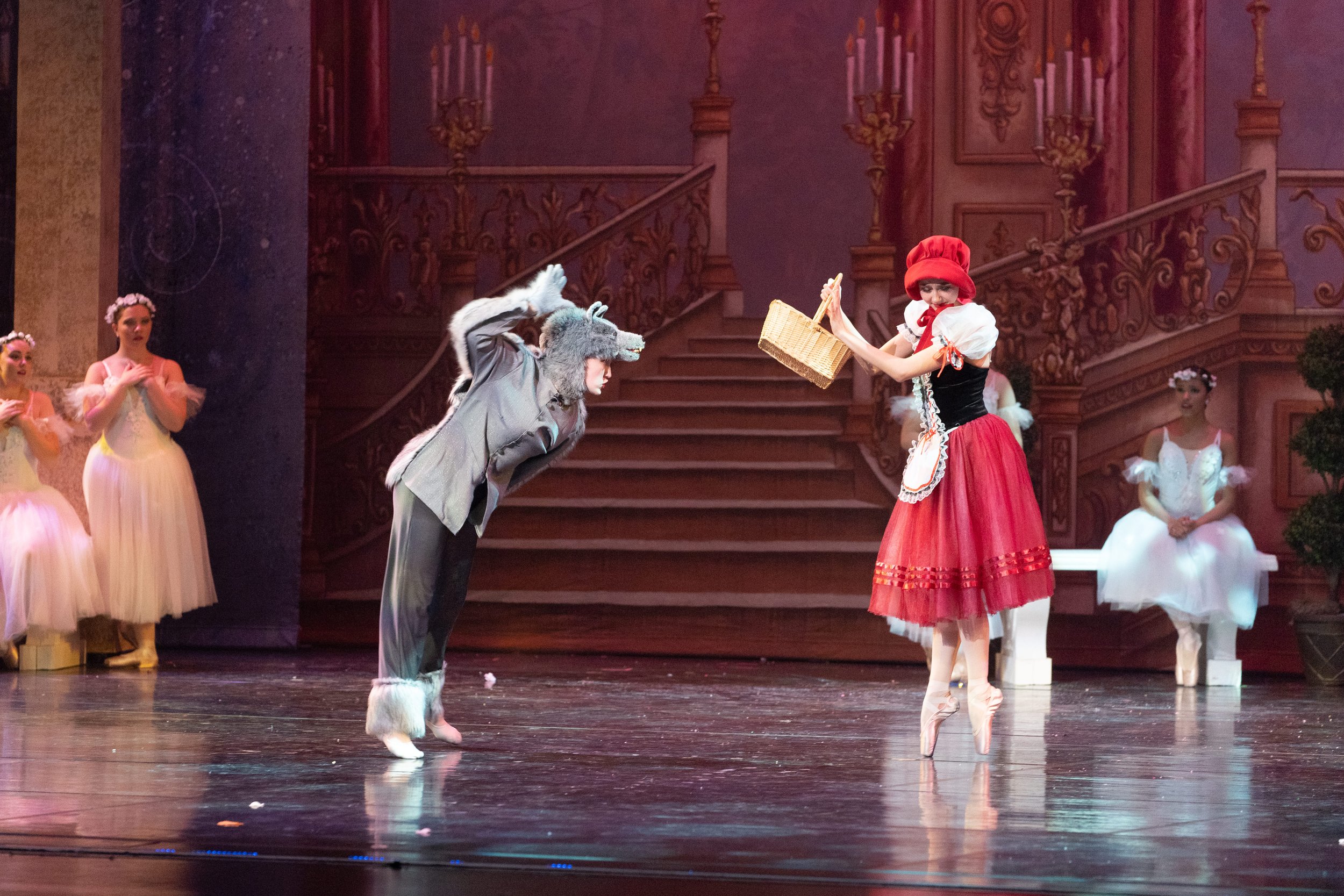 Ballet dancers performing on stage, with one dressed as a wolf in a gray costume and another as Little Red Riding Hood in a red skirt and bonnet, in a scene from a ballet production.