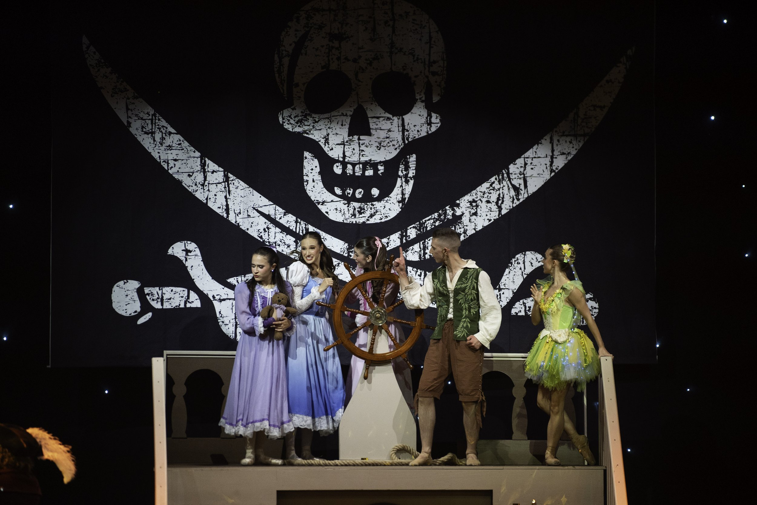 A theatrical performance of Peter Pan featuring five actors on stage, with a large skull and crossbones backdrop.