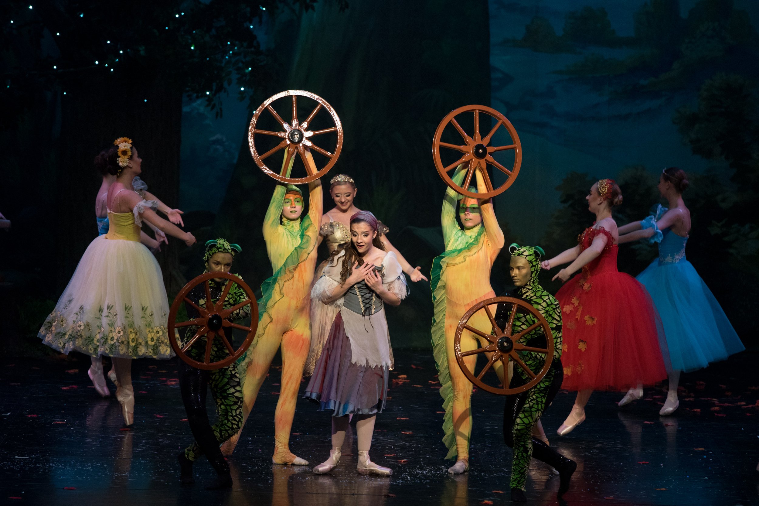 A ballet performance featuring dancers in colorful costumes, including women in flower and sky-colored dresses, and others dressed as animals with green and black patterns, with some holding large wooden wheels, on a stage with a painted forest backg