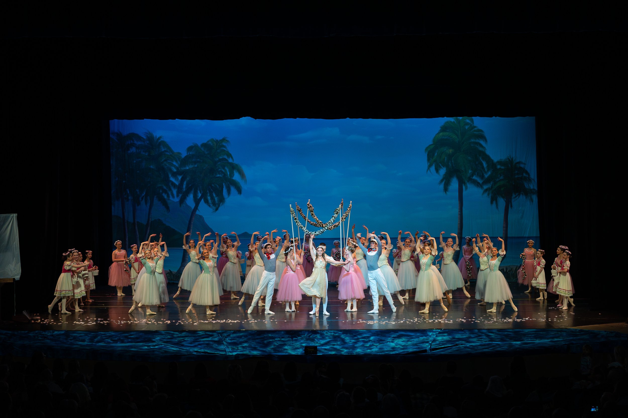 Ballet dancers on stage performing under a tropical backdrop with palm trees and blue sky, with some dancers in pink tutus and others in pastel dresses, during a scenic production.