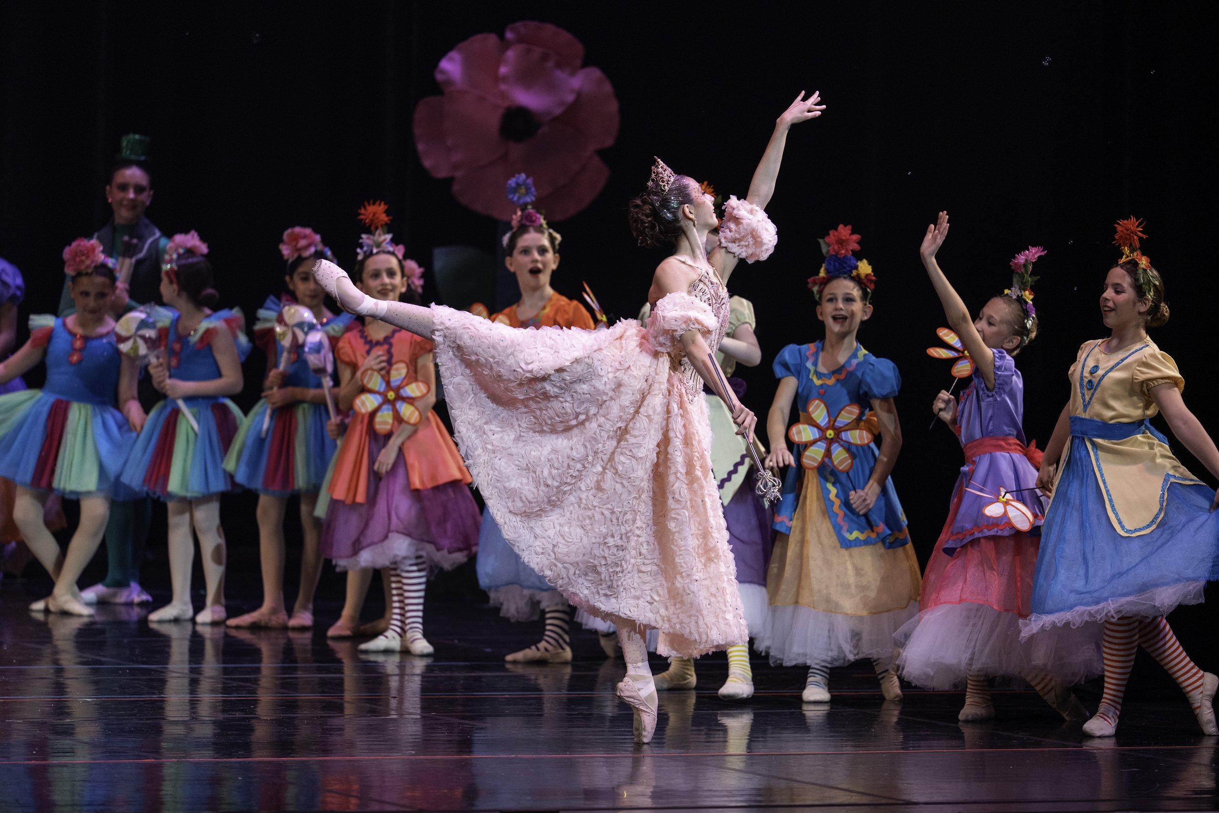 Children and an adult dressed in colorful costumes performing a ballet on stage. The background features a giant flower. The main dancer, a woman in a pink dress and tiara, is holding one leg up and extending one arm outward.