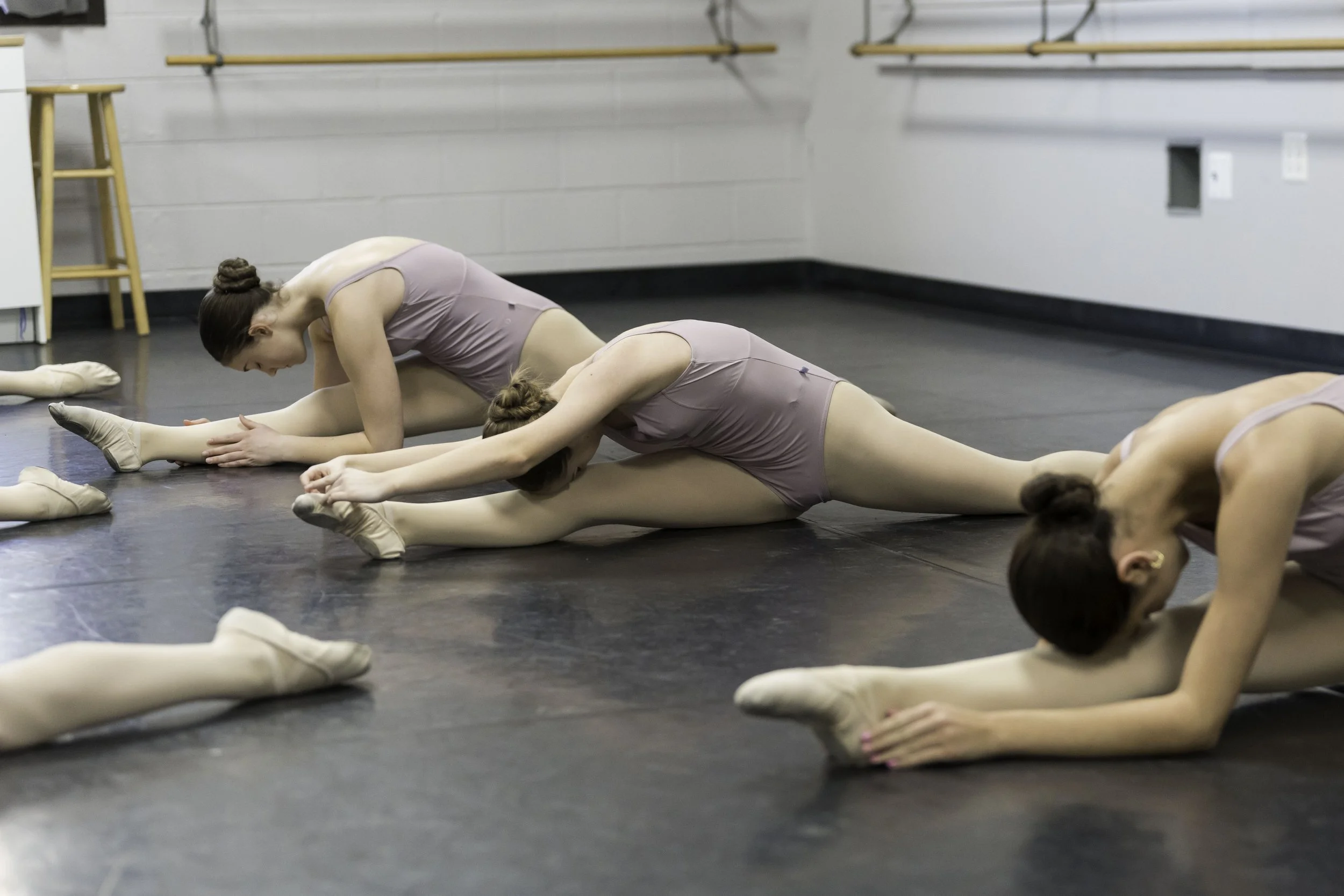 Ballet dancers in purple leotards and tights stretching on the floor in a dance studio