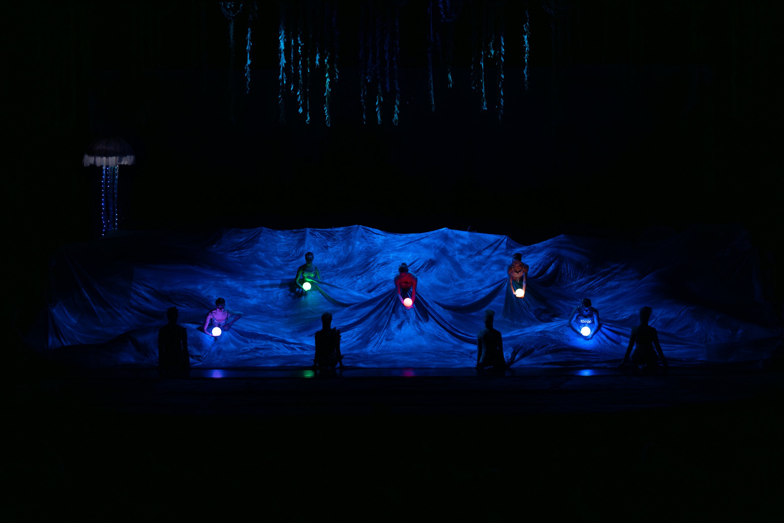 A stage performance with six performers holding glowing orbs, beneath hanging decorations resembling jellyfish, with a water-like textured backdrop illuminated in blue light.